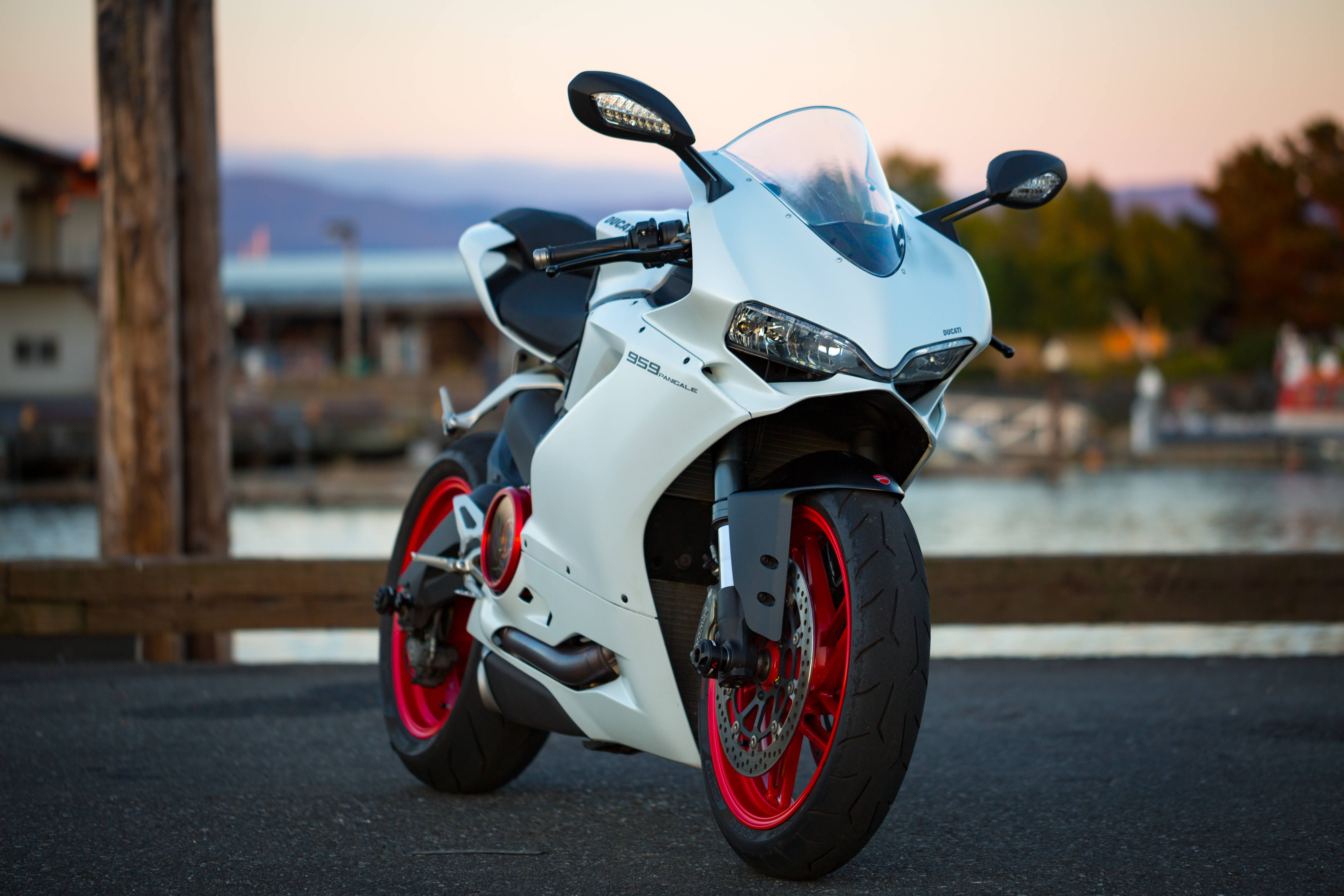 機車 桌布 壁紙 — 一架白色Ducati 959 Panigale電單車，紅色車輪，停泊在日落時分近水的瀝青路上。