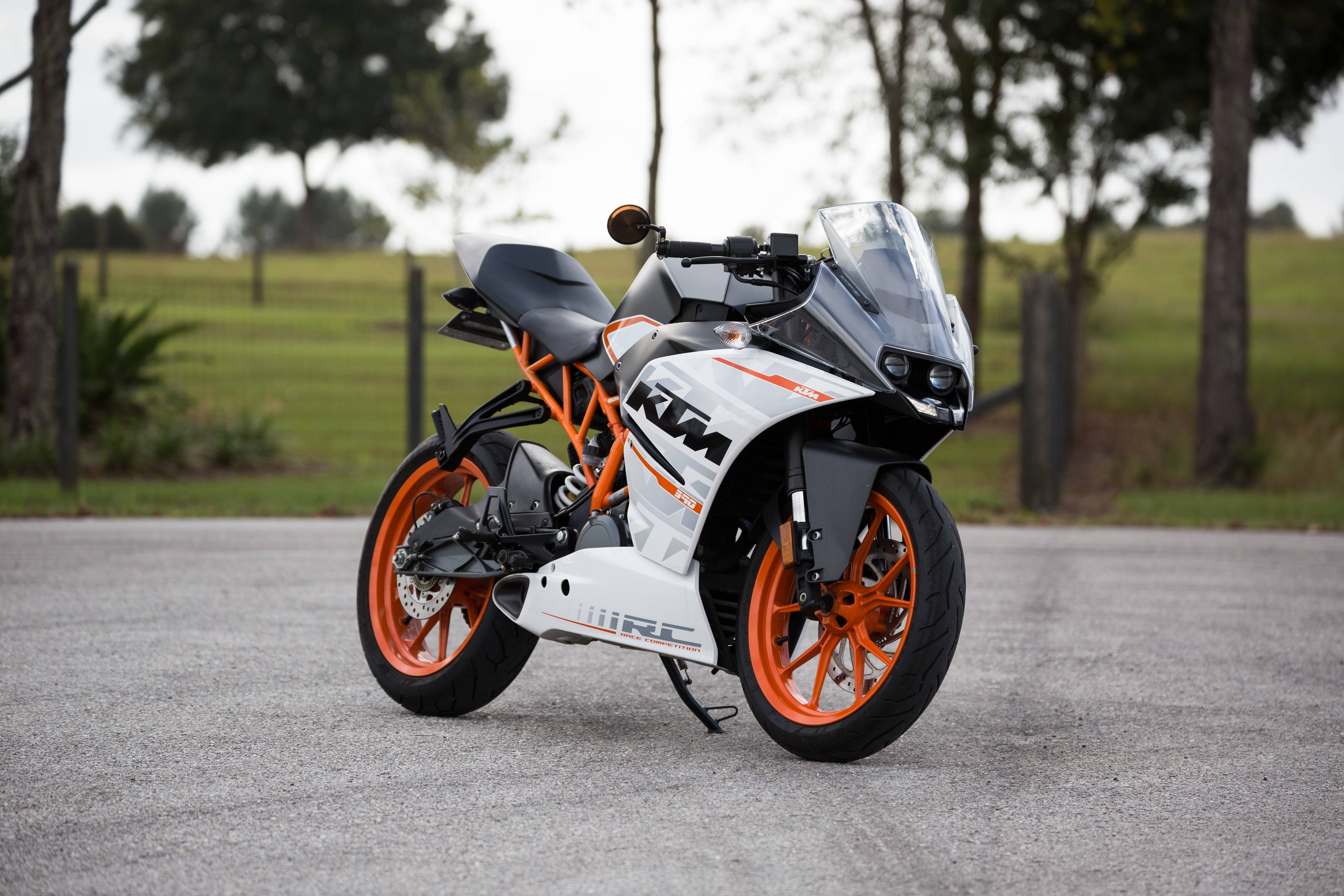 機車 桌布 壁紙 — 一架白色和橙色嘅KTM RC電單車，型格咁停泊喺碎石路上，背景有樹木同草地。