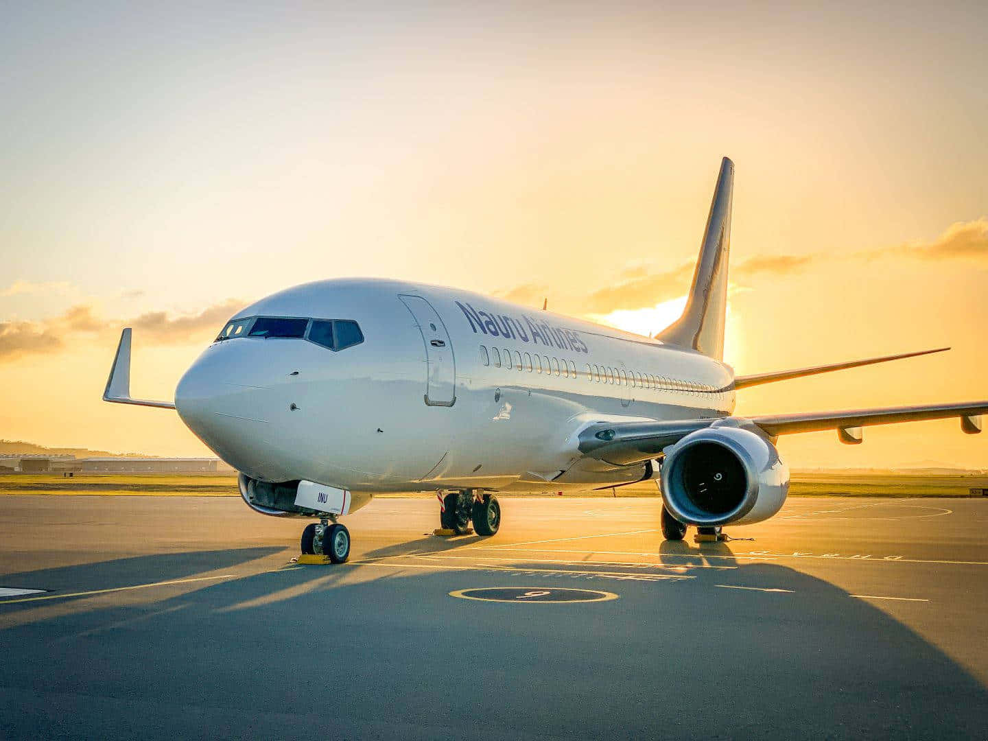 天橋 桌布 壁紙 — 一架白色的瑙魯航空客機停在機場停機坪上，沐浴在金色的夕陽光中，投下長長的影子。