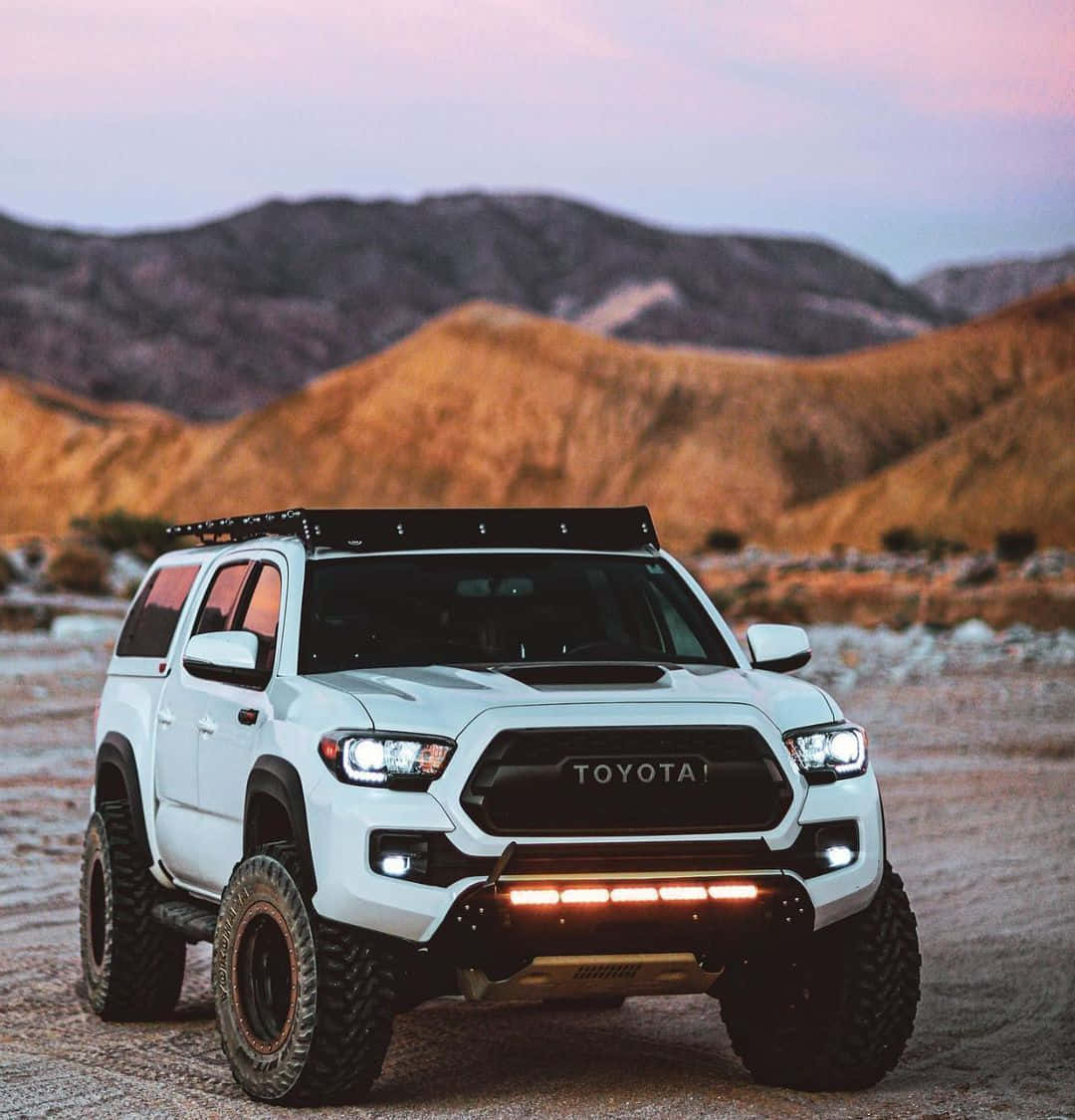 Off-road Car 桌布 壁紙 — 一架白色豐田 Tacoma 配備越野裝備，在日落時分準備好停在一條塵土飛揚的小徑上，背景是連綿起伏的沙漠山丘。