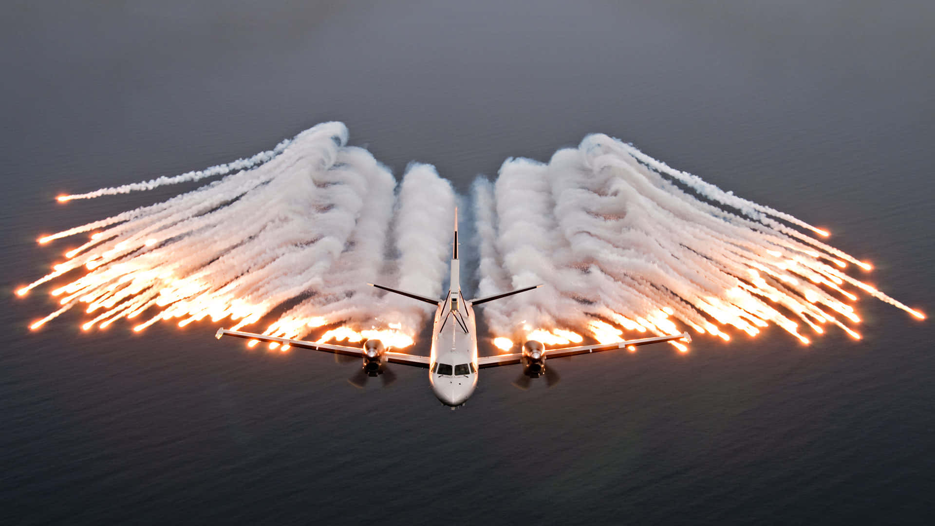 Air Force 桌布 壁紙 — 一架白色飛機在黑暗的水面上飛行，身後有火焰般的排氣軌跡形成天使翅膀。