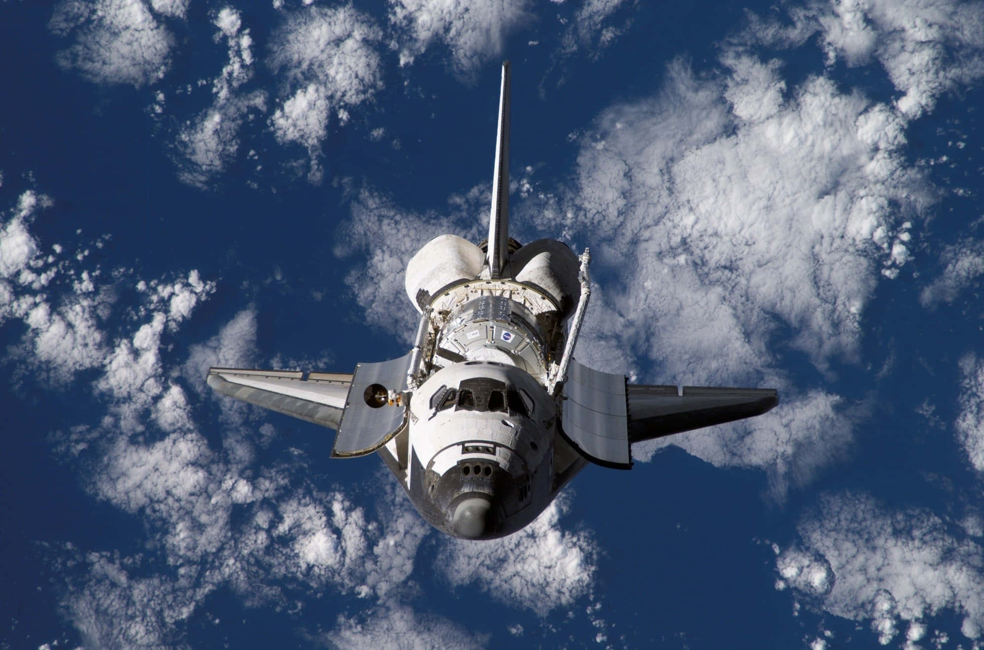 Space Mission 桌布 壁紙 — 一架穿梭機在地球上空翱翔，蓬鬆的白雲在其機翼下飄動，襯托著深藍色的天空。