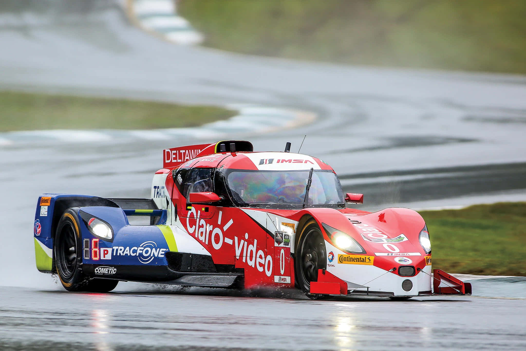 Car Racing 桌布 壁紙 — 一架紅白藍賽車在濕滑的彎道上飛馳，賽道被雨水浸透，兩旁是草坡。