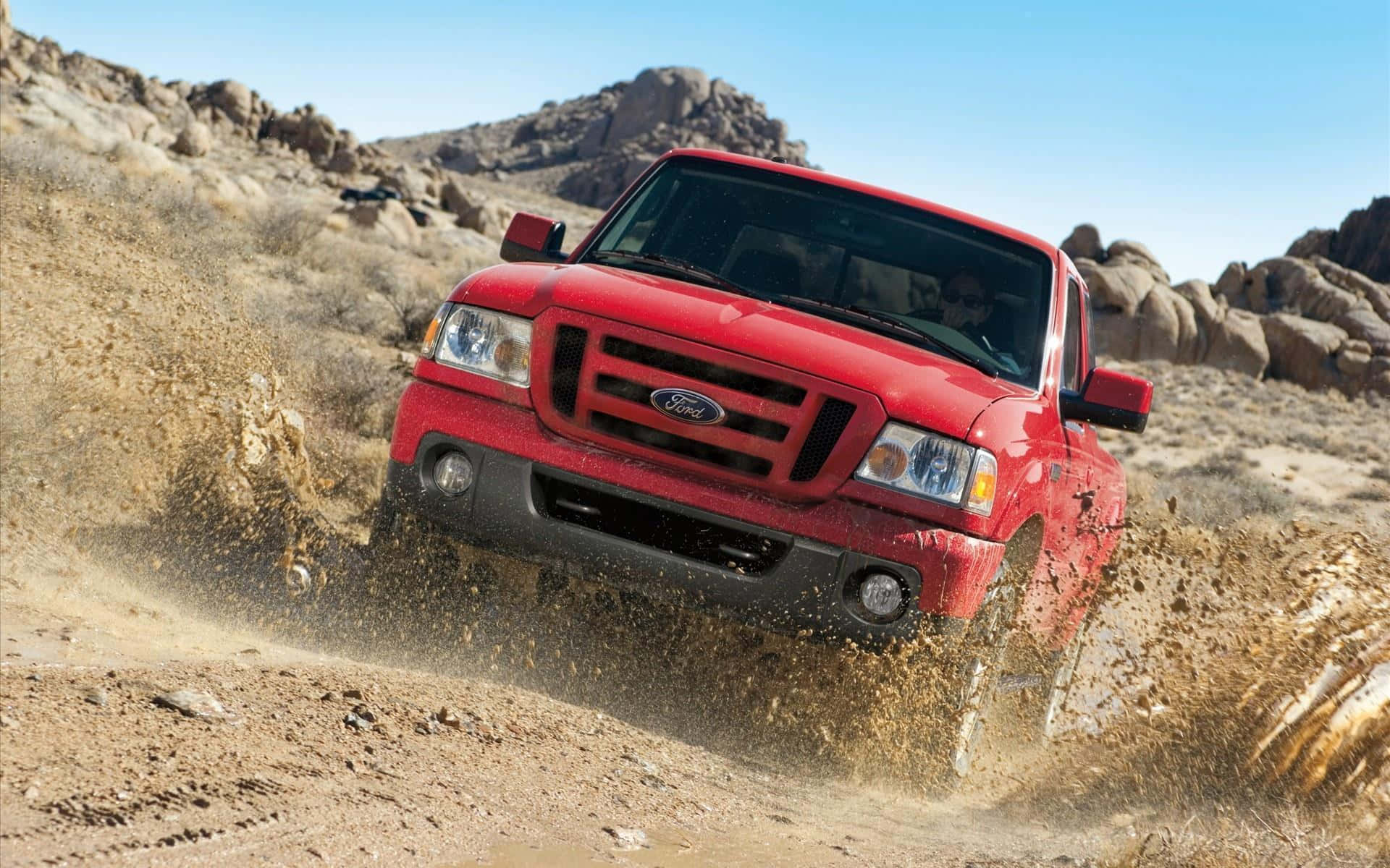 Ford Ranger 桌布 壁紙 — 一架紅色福特貨車在晴朗藍天下，沿著塵土飛揚的沙漠小徑行駛，揚起沙塵撲向岩石丘陵。