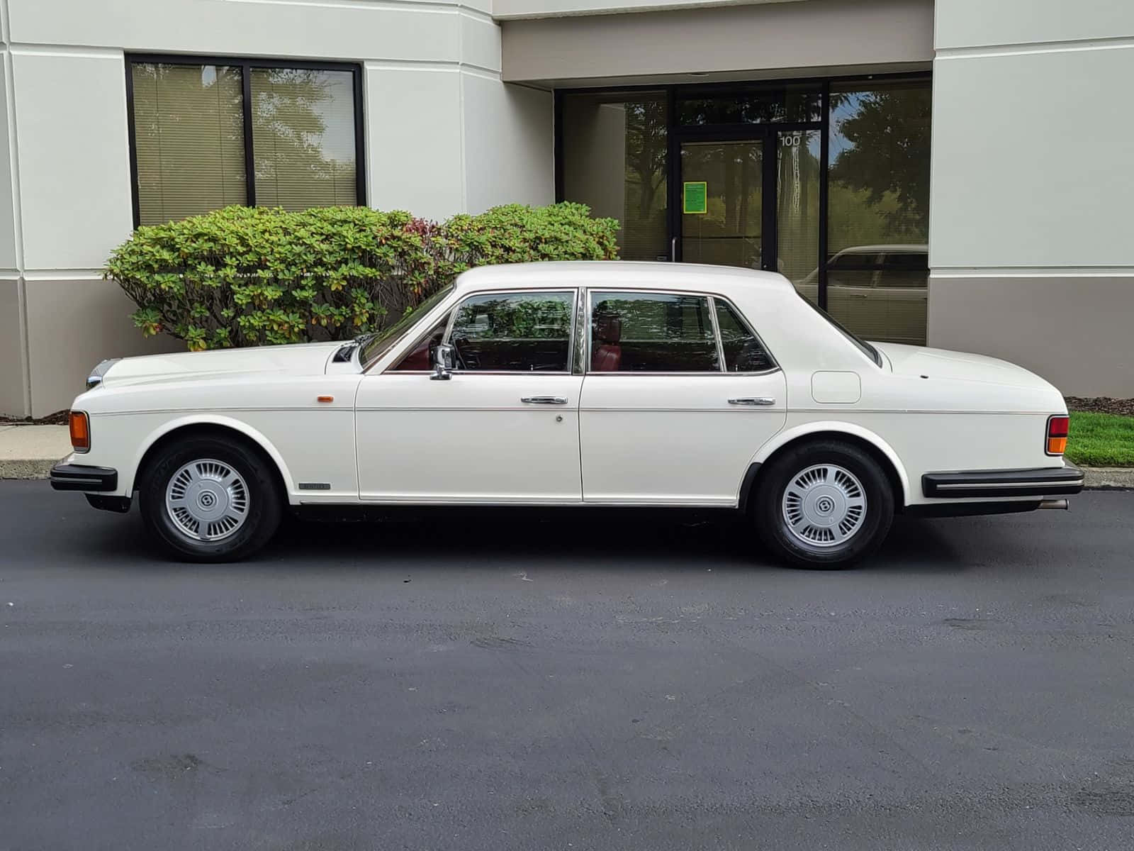 Bentley Eight 桌布 壁紙 — 一架純白色勞斯萊斯 Silver Shadow II 停泊在現代建築物前，旁邊有綠色灌木。