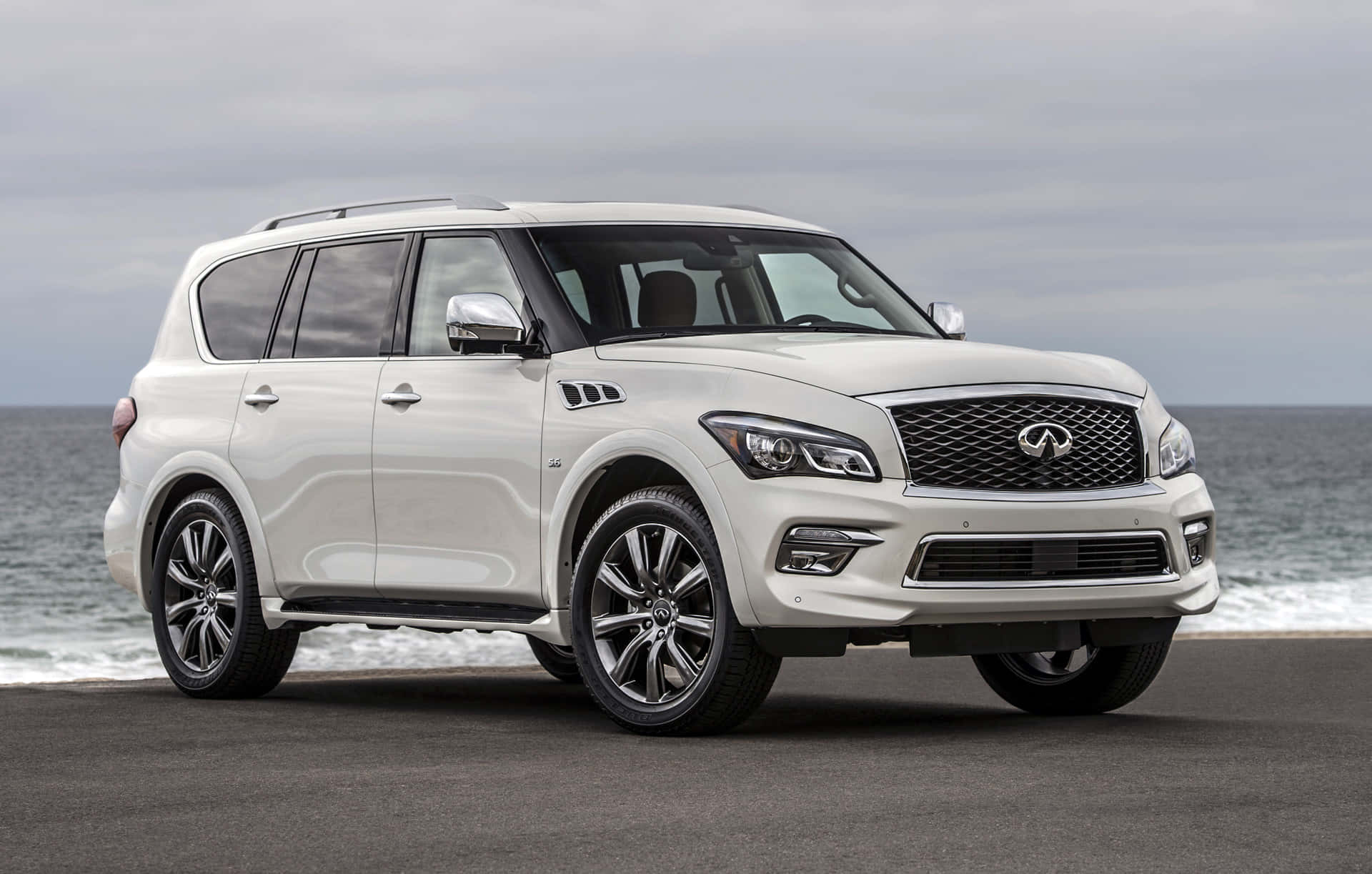 Infiniti Qx80 桌布 壁紙 — 一架純白色的 Infiniti QX80 SUV 停泊在海岸公路上，背後有海浪拍打，天空多雲。