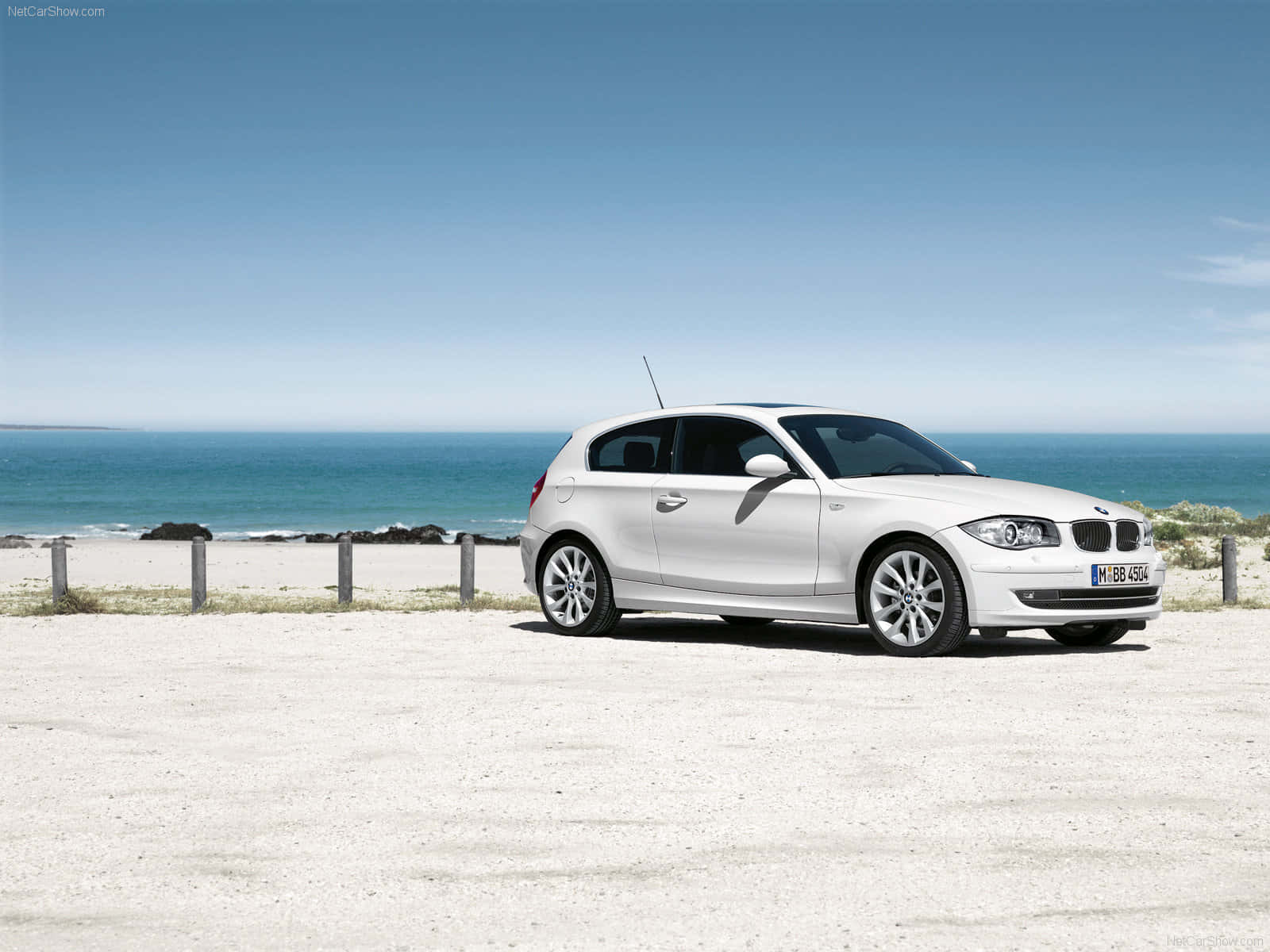 Bmw 1 Series 桌布 壁紙 — 一架純白色的BMW 1系掀背車停泊在陽光普照的海岸碎石停車場，背景是海洋和天空。