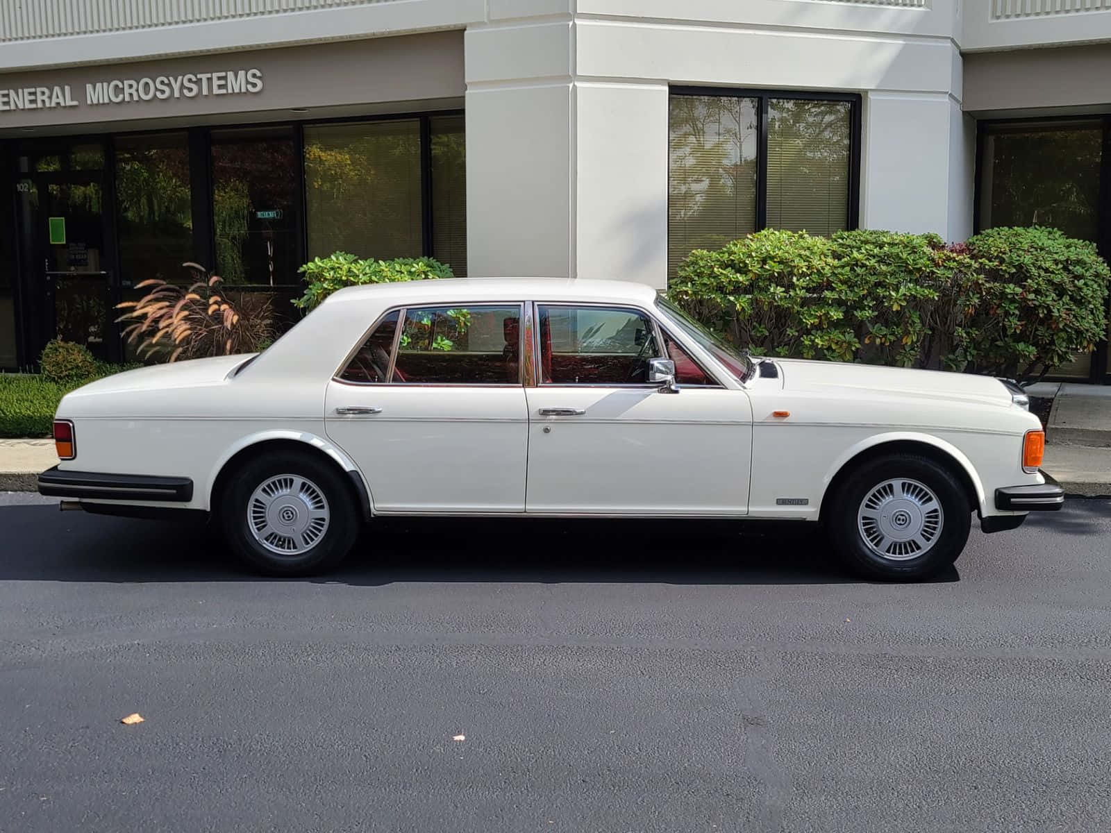 Bentley Eight 桌布 壁紙 — 一架純白色的勞斯萊斯銀影 II 停泊在現代辦公大樓前的瀝青路上，周圍有綠色灌木。