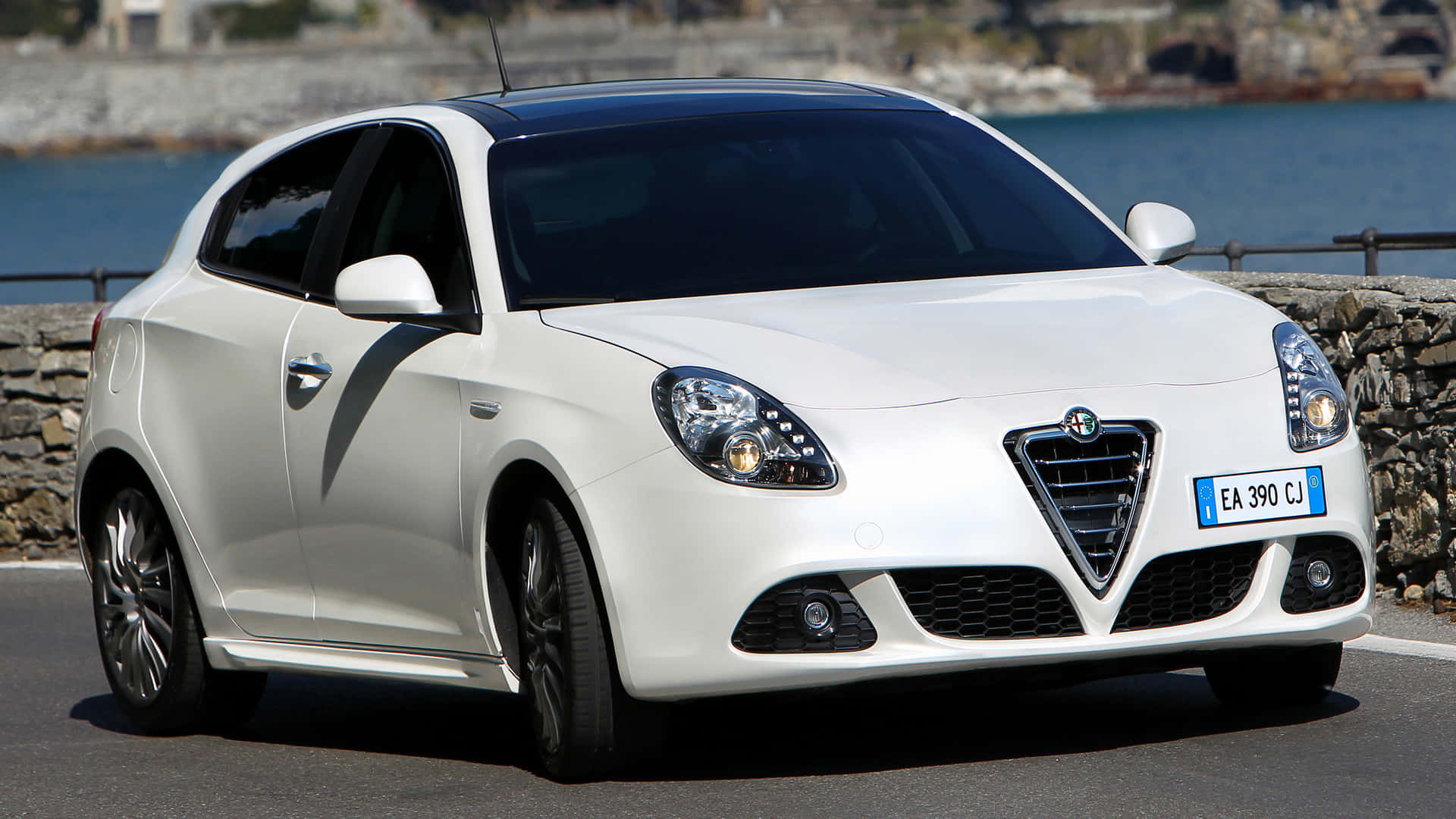Alfa Romeo Giulietta 桌布 壁紙 — 一架純白色的愛快羅密歐Giulietta停泊在沿海道路上，背後有石牆和藍色大海。