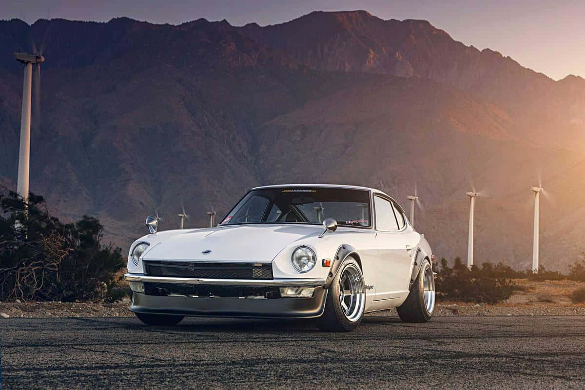 Datsun 桌布 壁紙 — 一架純白色經典跑車停泊在瀝青路上，背景是風力發電機和崎嶇的山脈，沐浴在金黃色的日落光芒中。