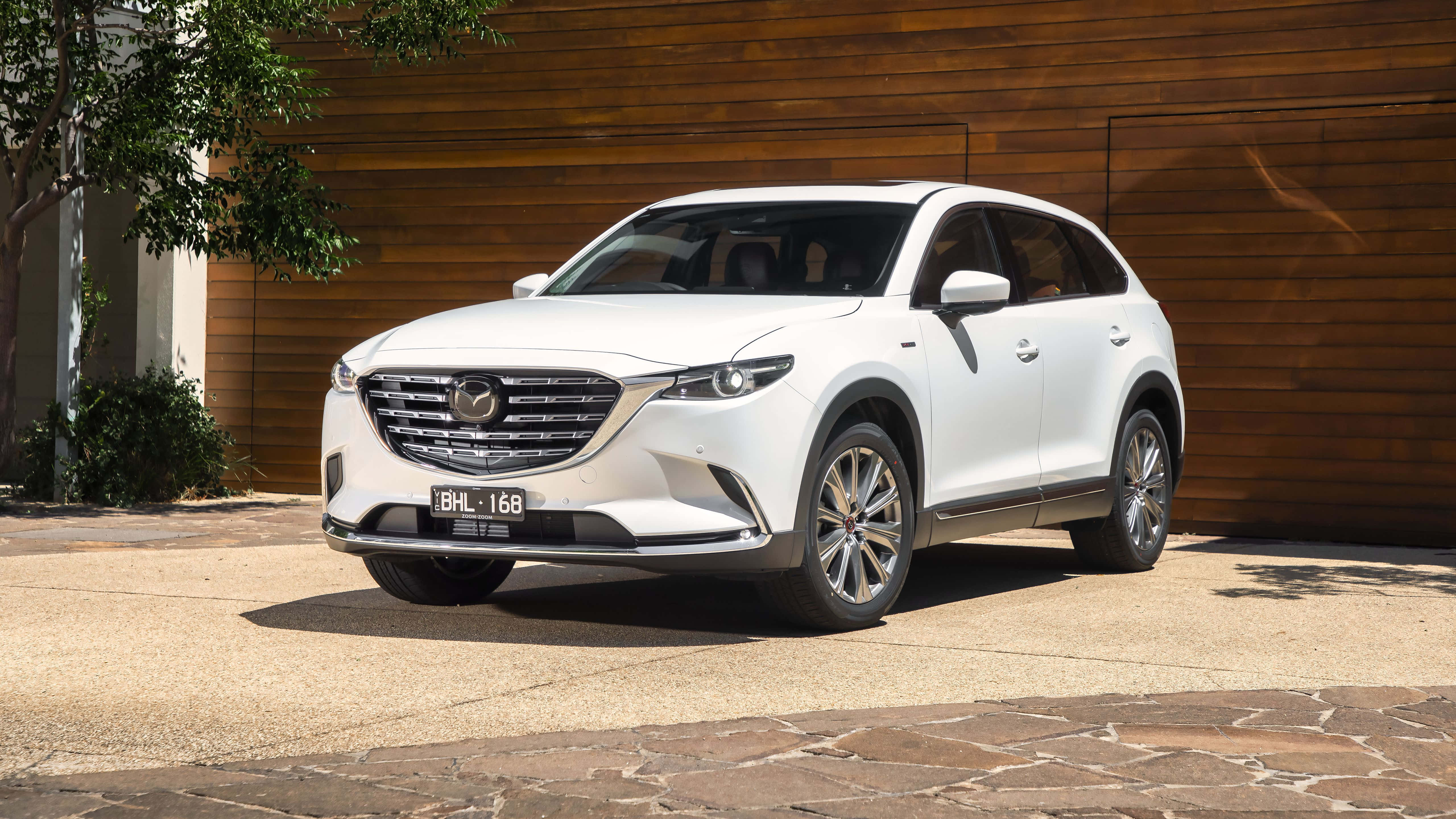Mazda Cx-9 桌布 壁紙 — 一架純白色萬事得CX-90 SUV停泊在石頭車道上，旁邊是木牆，在明亮的日光下。
