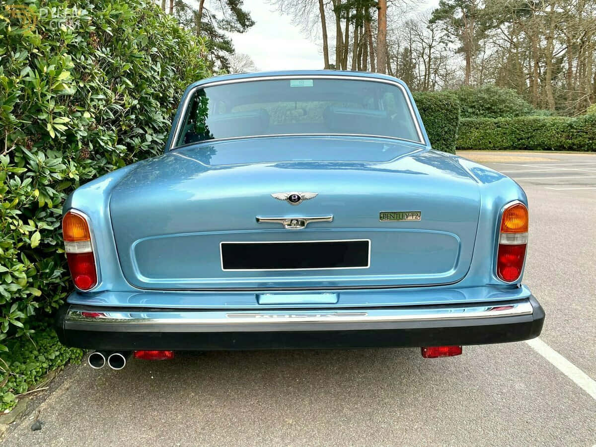 Bentley T2 桌布 壁紙 — 一架經典淺藍色 Bentley T2 停泊在瀝青路上，旁邊有綠色籬笆，天空多雲。