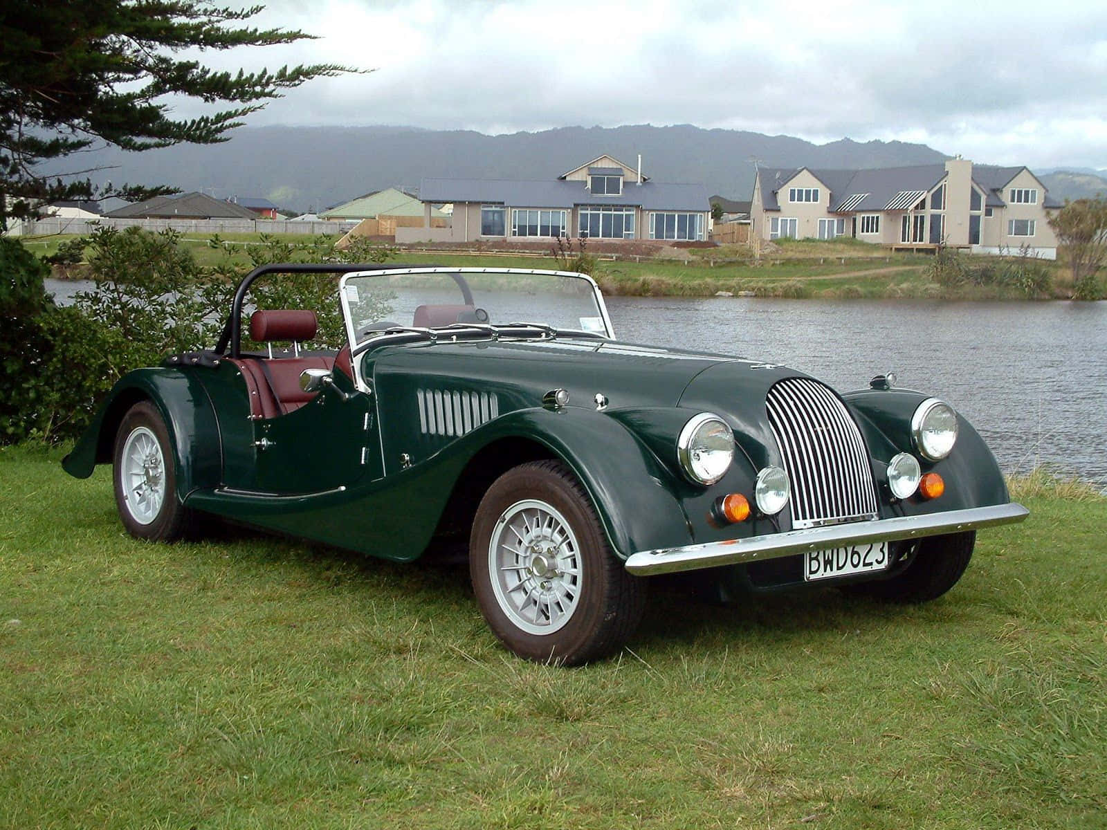 Morgan 桌布 壁紙 — 一架經典綠色摩根開篷跑車，配紅色皮革座椅，停泊在湖邊草地上，背景有房屋和山丘，天空多雲。