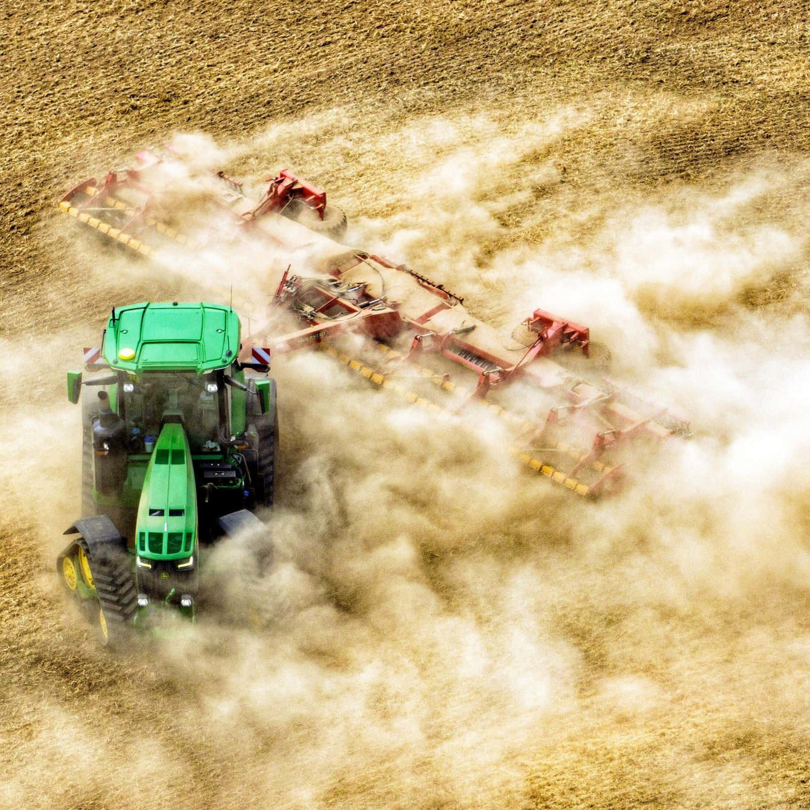 John Deere 桌布 壁紙 — 一架綠色拖拉機拖著紅色犁具穿過乾燥土壤，揚起灰塵的航拍畫面。