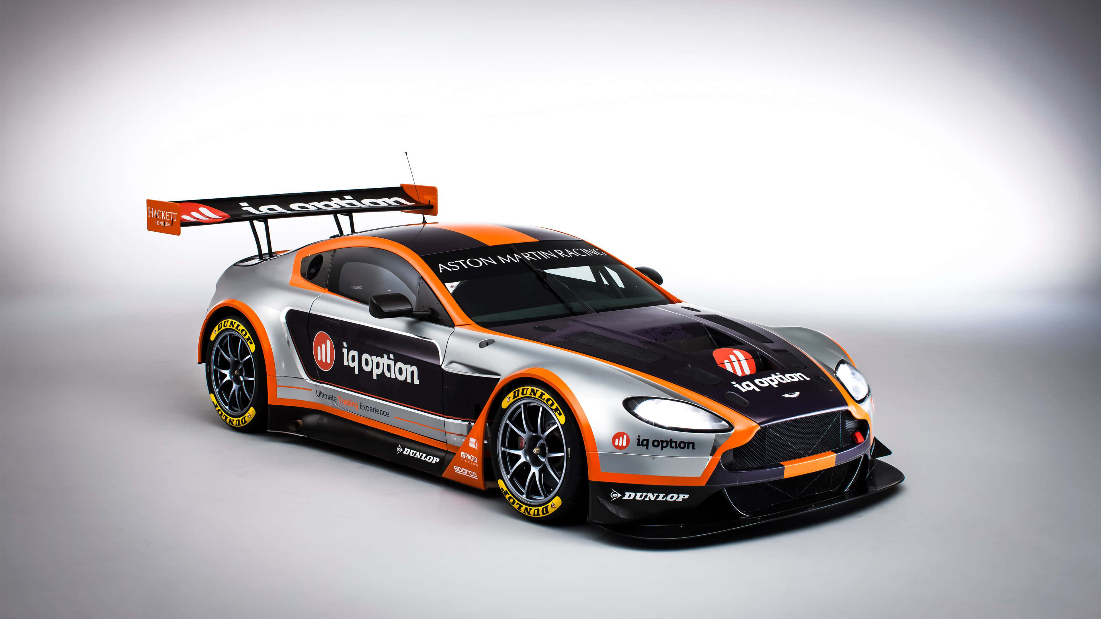 Car Racing 桌布 壁紙 — 一架線條流暢的Aston Martin賽車，採用橙色和黑色塗裝，在漸變灰色工作室背景前蓄勢待發。