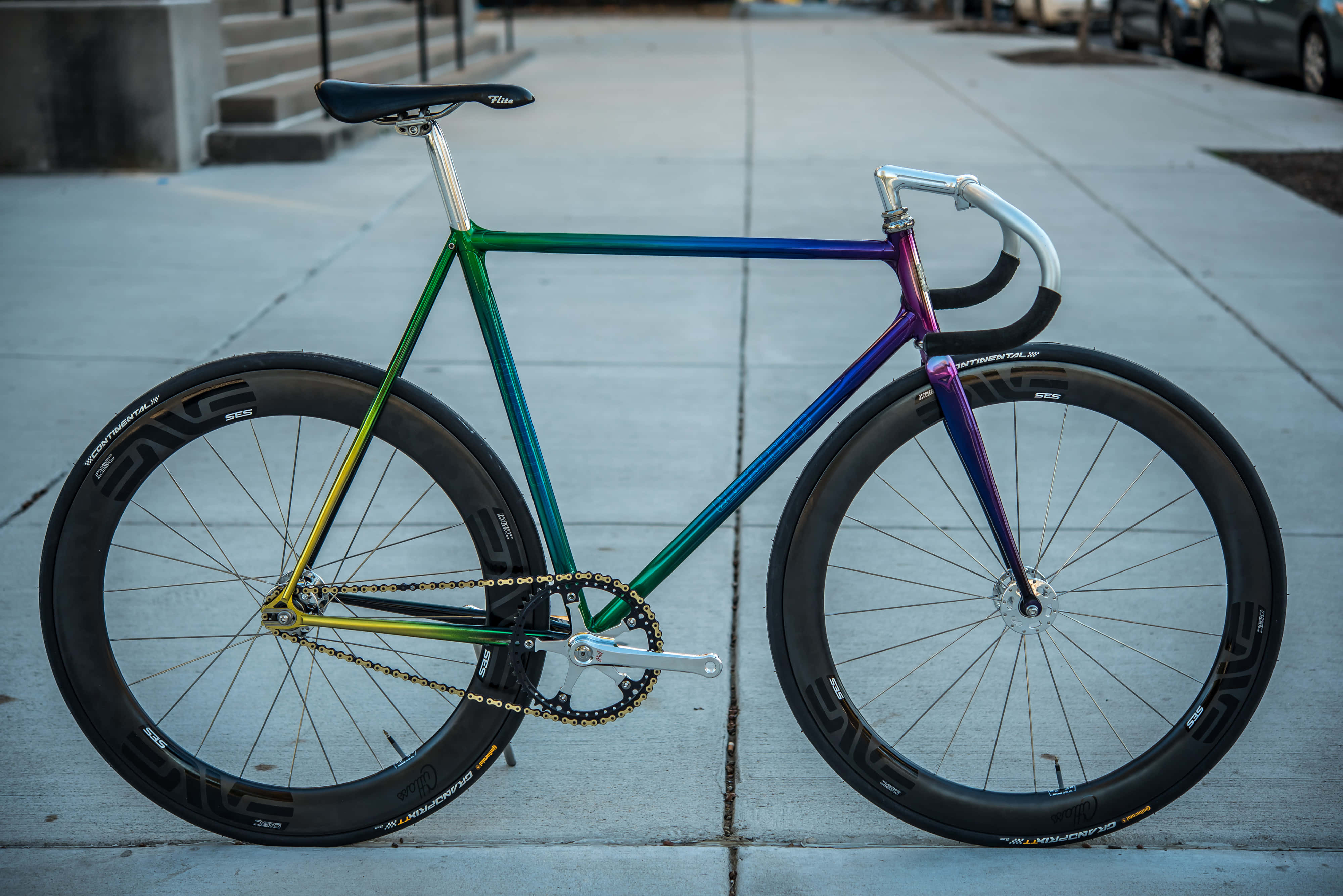 Bicycle 桌布 壁紙 — 一架色彩繽紛的彩虹色固定齒輪單車佇立在混凝土行人路上，其流線型車架在柔和日光下閃閃發光。