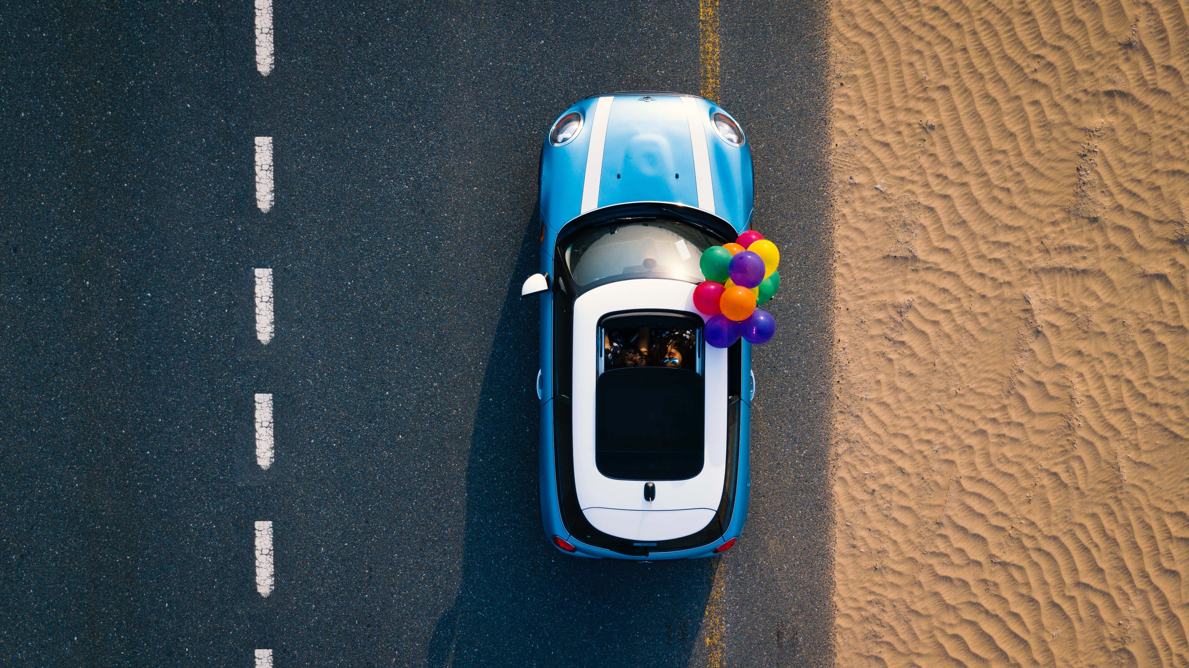 road trip 桌布 壁紙 — 一架藍白色汽車載著彩色氣球，行駛在波紋沙灘旁邊的道路上的鳥瞰圖。