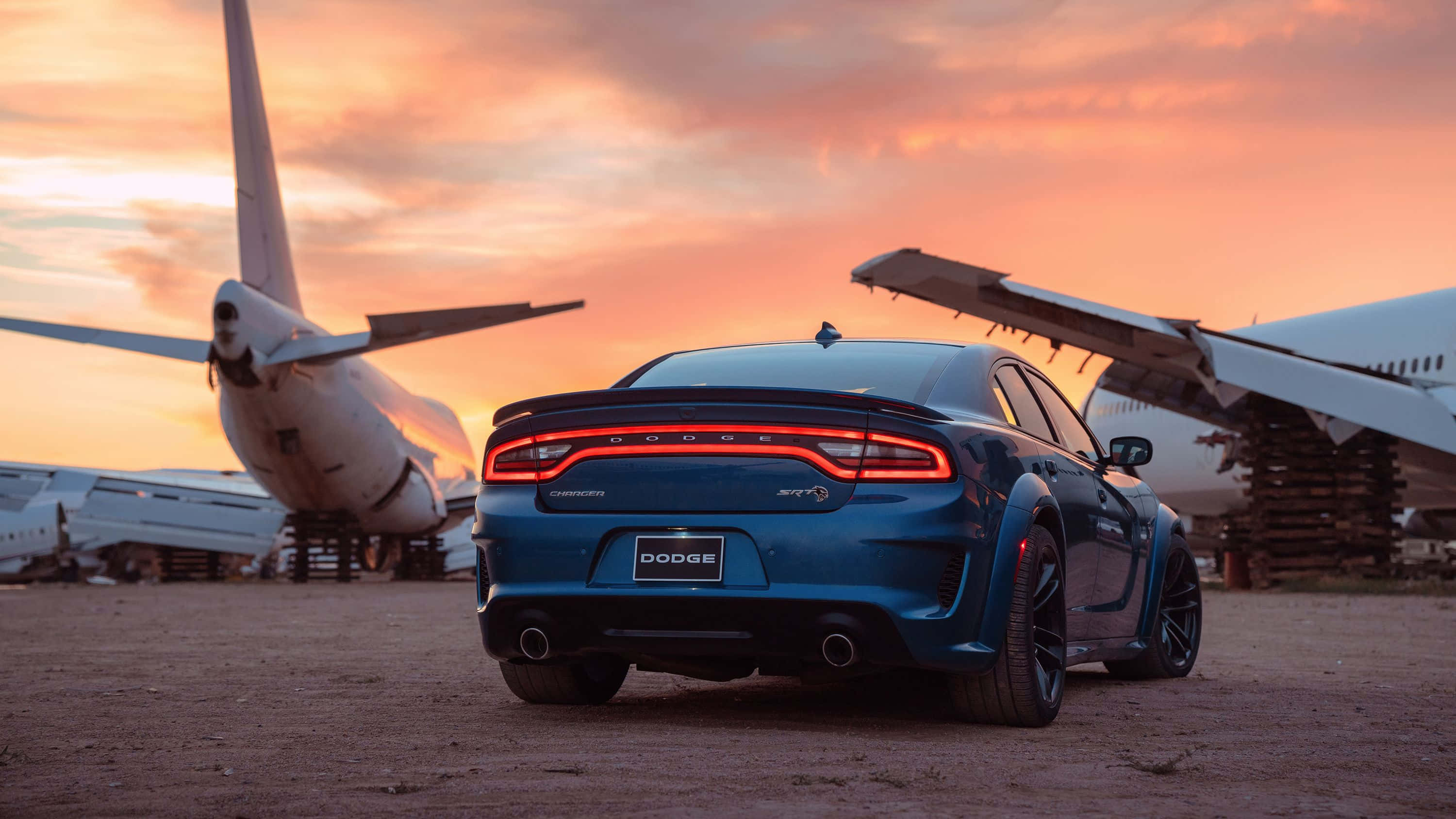 Dodge Charger 桌布 壁紙 — 一架藍色Dodge Charger SRT停泊在兩架退役飛機之間，背景是熾熱的日落天空。