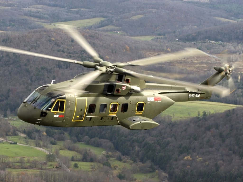 Helicopter 桌布 壁紙 — 一架軍用直升機低空飛過起伏的森林景觀，下方有綠色田野和蜿蜒的道路。