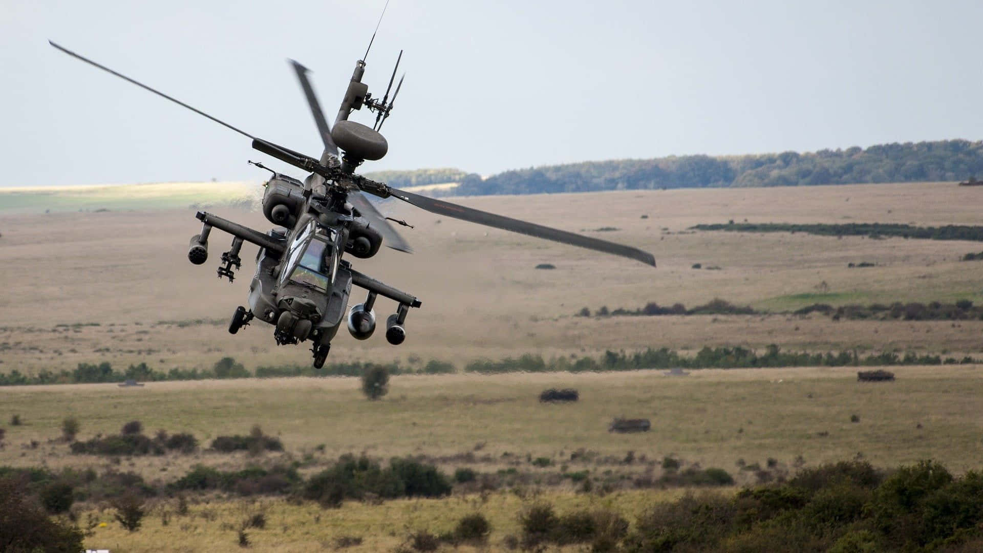 Helicopter 桌布 壁紙 — 一架軍用直升機在廣闊的開闊田野上空低空盤旋，遠處有樹線，天空晴朗。