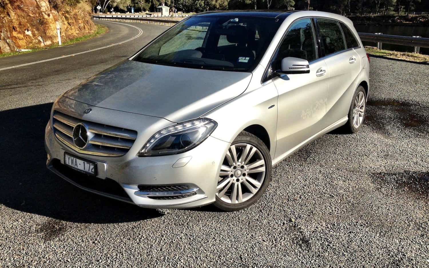 Mercedes Benz B-class 桌布 壁紙 — 一架銀色 Mercedes-Benz 旅行車停泊在碎石路上，旁邊是蜿蜒的道路，背景有樹木和岩石峭壁。