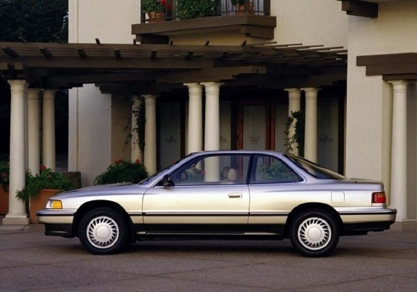 Acura Legend 桌布 壁紙 — 一架銀色1990年代房車停泊在柱式涼棚下，前方係米色灰泥建築物，配有盆栽植物。
