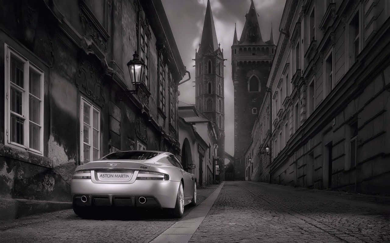 Black And White Car 桌布 壁紙 — 一架銀色Aston Martin跑車停泊在鵝卵石街道上，兩旁是帶有哥德式尖頂的古老歐洲建築，天空陰沉多雲。