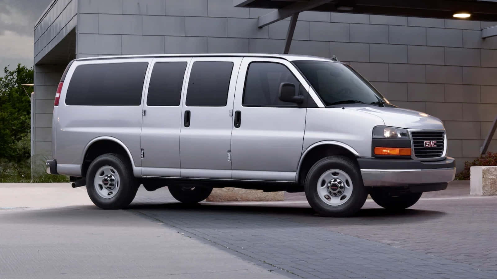 Gmc Savana 桌布 壁紙 — 一架銀色GMC客貨車停泊在鋪砌的停車場上，旁邊是一座現代灰色磚砌建築，天空多雲。