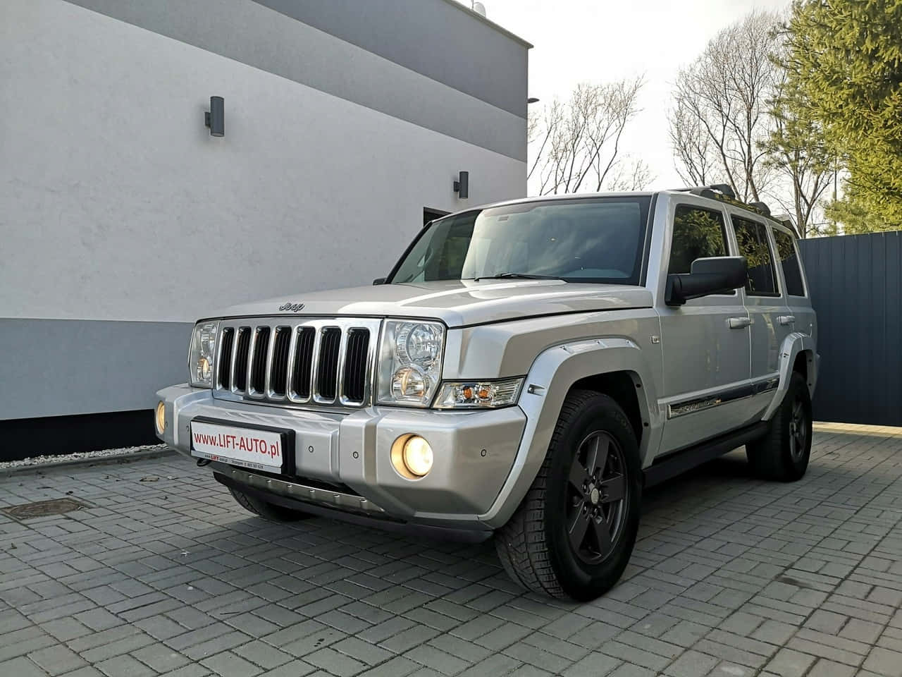 Jeep Commander 桌布 壁紙 — 一架銀色Jeep Commander SUV停泊在灰色磚砌車道上，旁邊是一座現代建築，背景有光禿禿的樹木。