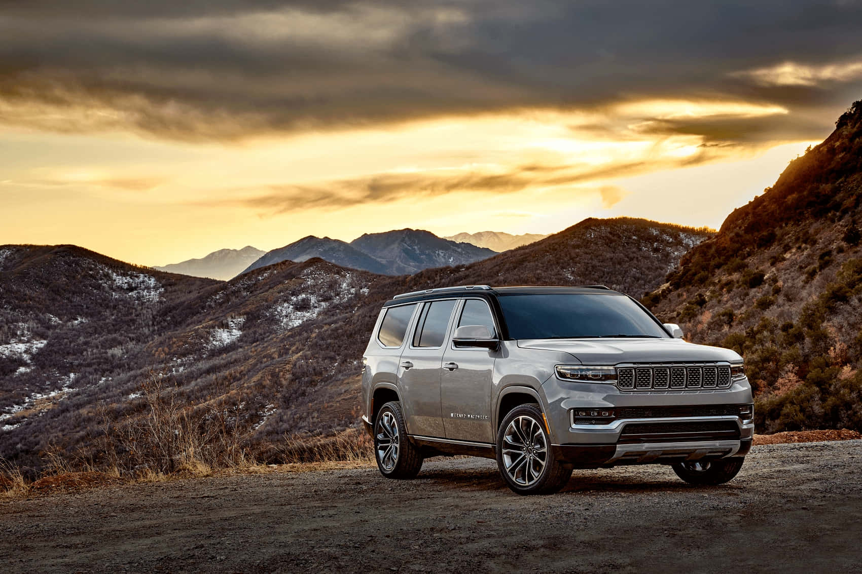 Jeep Wagoneer 桌布 壁紙 — 一架銀色Jeep Grand Wagoneer在日落時分停泊在泥路上，背景是積雪的山脈。