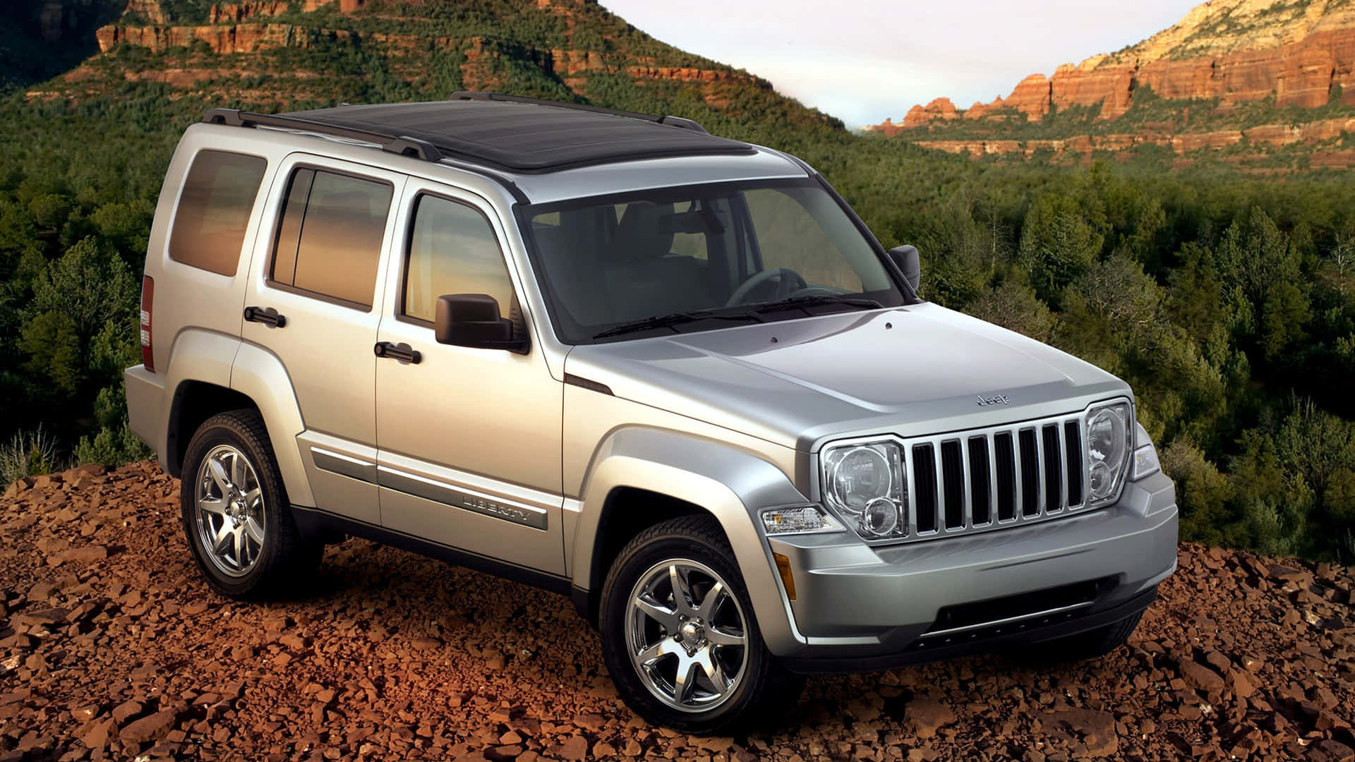 Jeep Liberty 桌布 壁紙 — 一架銀色Jeep Liberty SUV停泊在岩石地形上，背景有紅色岩層和綠色樹木，天空柔和。