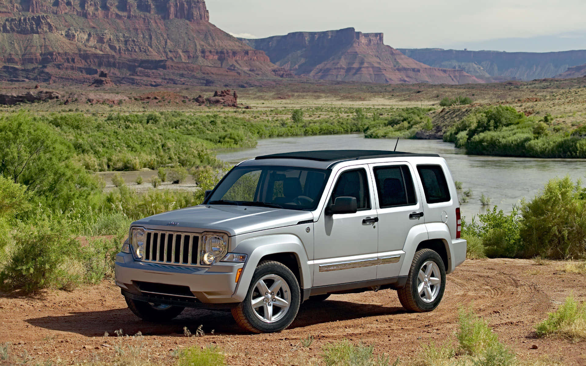 Jeep Liberty 桌布 壁紙 — 一架銀色Jeep Liberty停泊在河邊的泥路上，背景是紅色岩石峽谷懸崖。