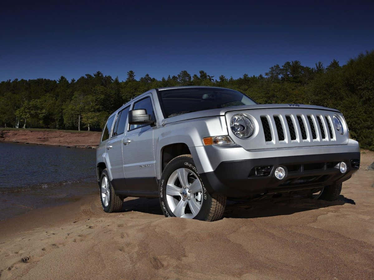 Jeep Patriot 桌布 壁紙 — 一架銀色Jeep Patriot SUV停泊在沙質岸邊，旁邊是平靜的水面，背後有茂密森林，天空清澈湛藍。