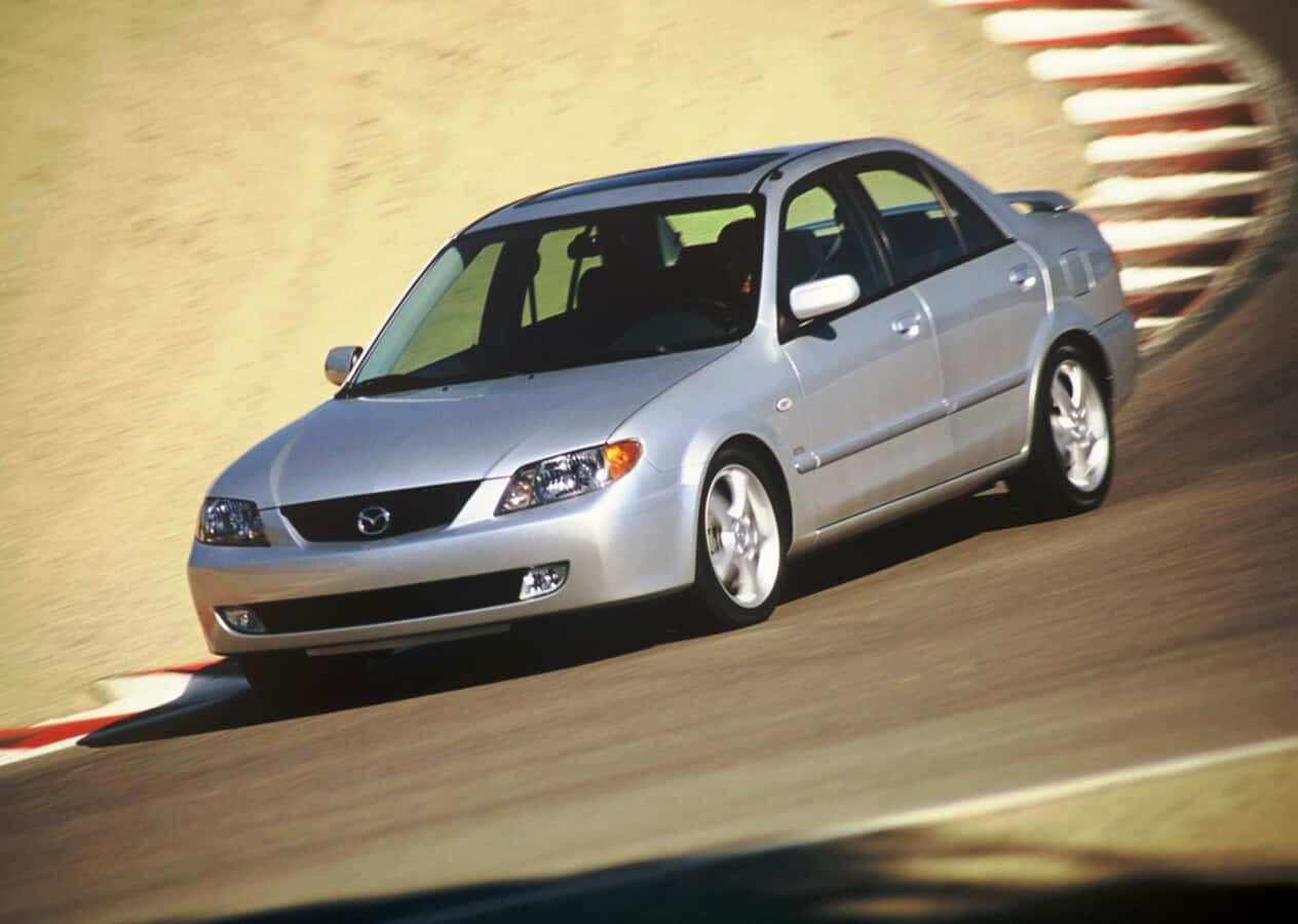 Mazda Protege 桌布 壁紙 — 一架銀色Mazda房車高速駛過賽車場彎道，賽道表面呈現動態模糊效果。