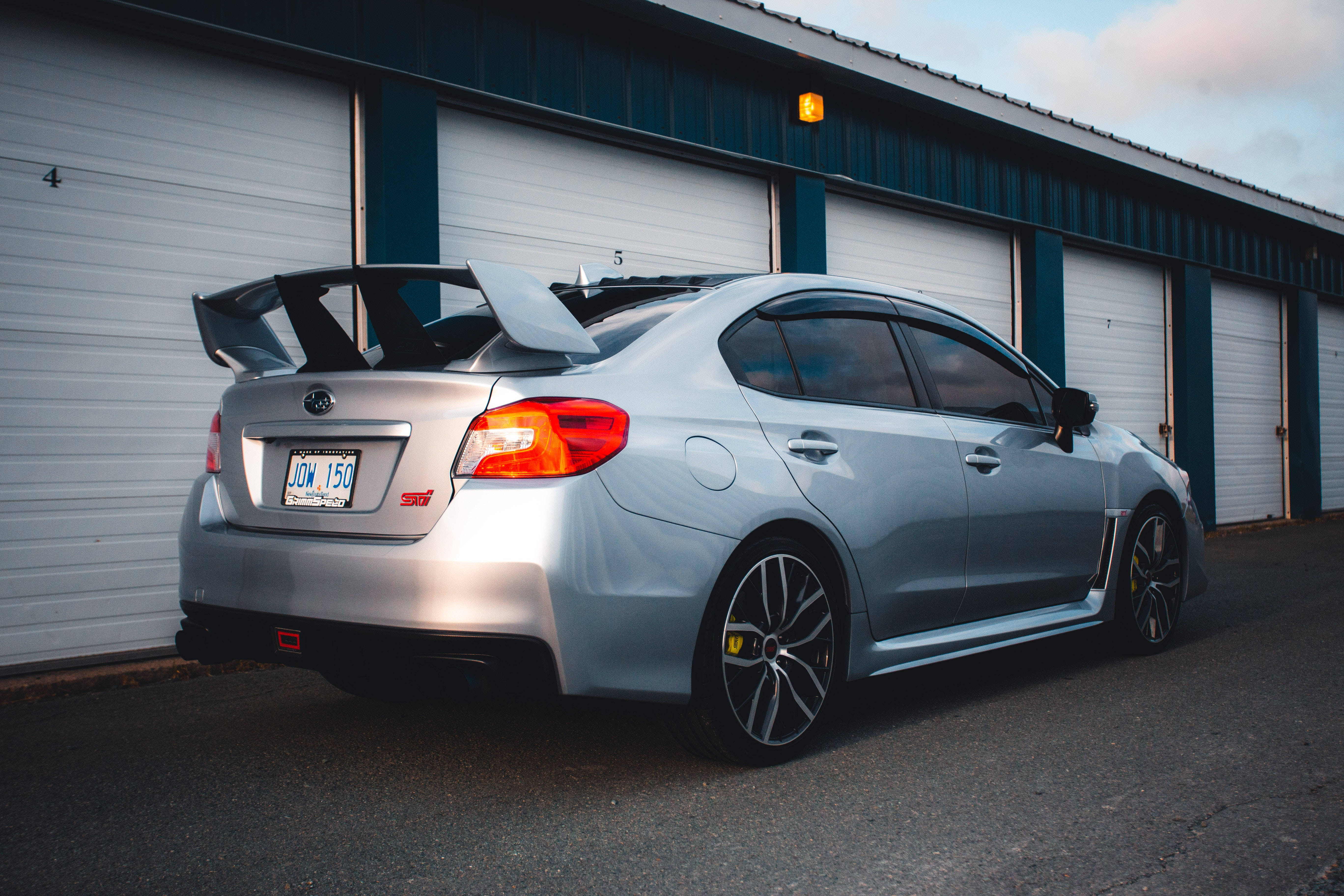 Sports Car 桌布 壁紙 — 一架銀色Subaru WRX STI配備大型尾翼，在黃昏時停泊在工業車庫門前。