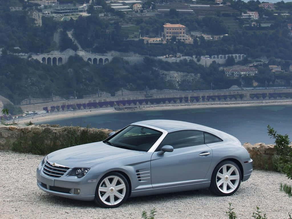 Chrysler Crossfire 桌布 壁紙 — 一架銀色克萊斯勒Crossfire跑車停泊在碎石觀景點，背景有沿海小鎮和拱橋。
