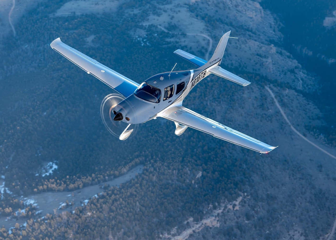 Aviation 桌布 壁紙 — 一架銀色單引擎螺旋槳飛機在崎嶇的山區地形上空翱翔，蜿蜒的道路和稀疏的樹木清晰可見。