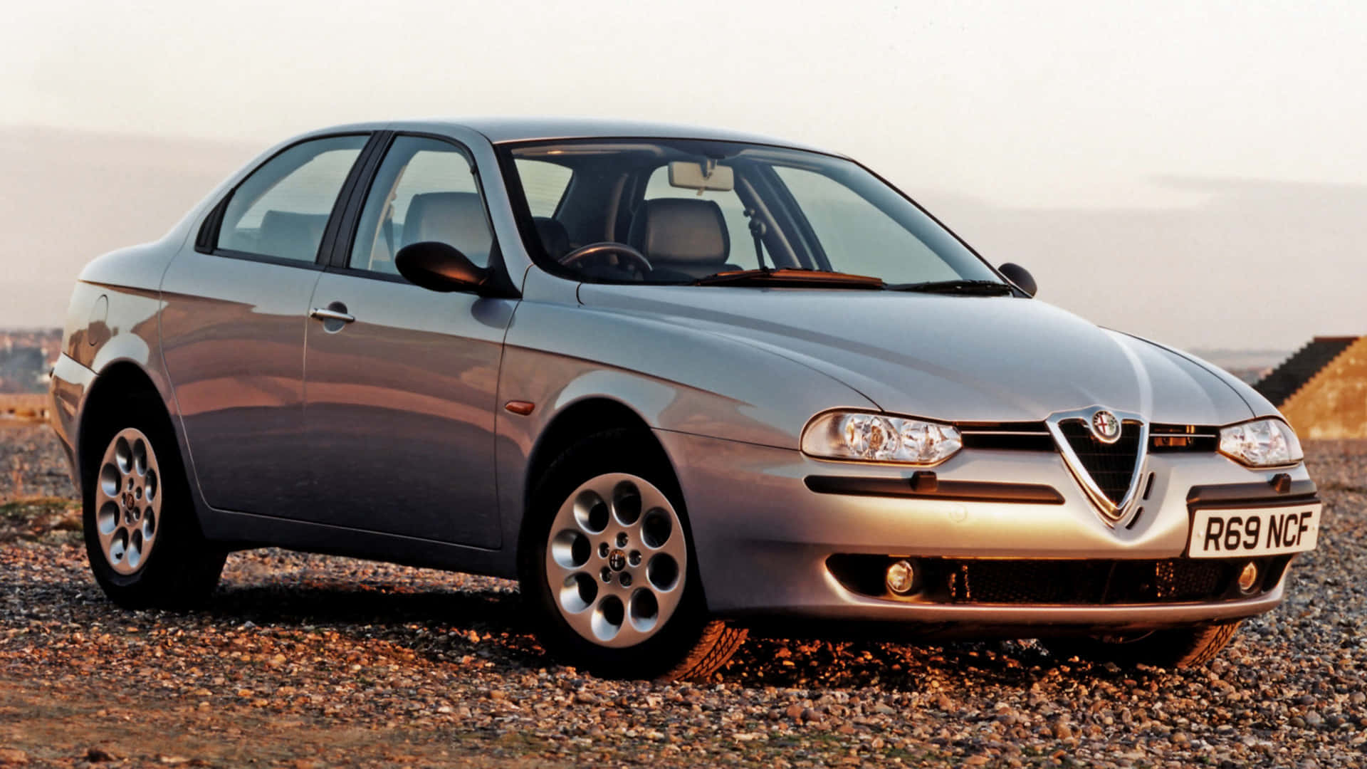 Alfa Romeo 156 桌布 壁紙 — 一架銀色嘅Alfa Romeo 156房車泊喺碎石場上，喺柔和嘅黃金時段光線下。