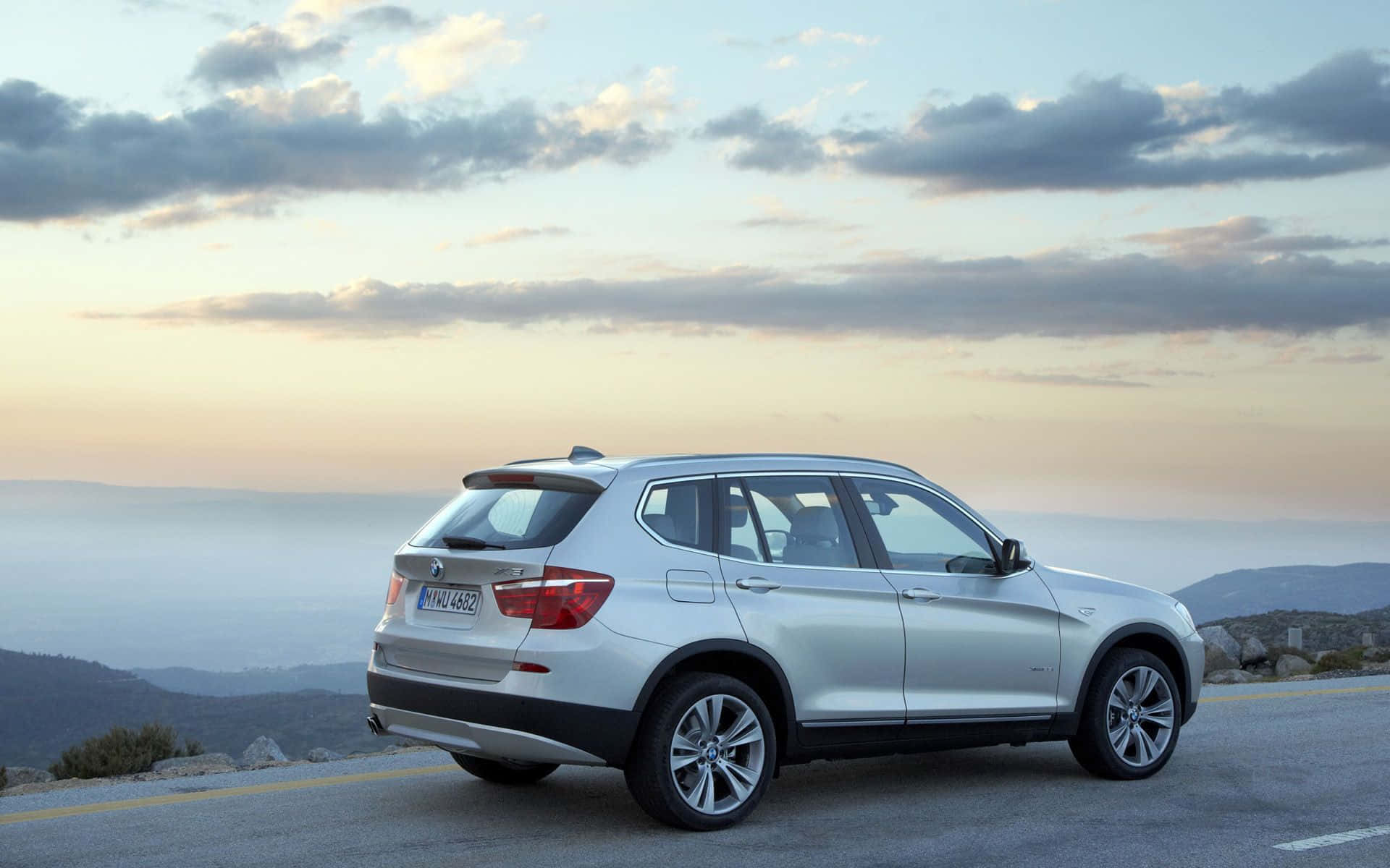 Bmw X3 桌布 壁紙 — 一架銀色寶馬SUV停泊在日落時分的海岸山路上，背景有戲劇性的雲層和遠山。