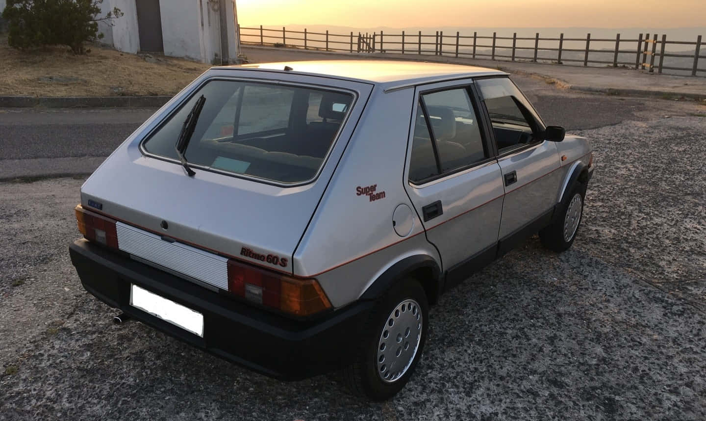 Fiat Ritmo 桌布 壁紙 — 一架銀色快意 Ritmo 60S 揭背車停泊在日落時分的碎石路上，背景有木欄柵和遠方的大海。
