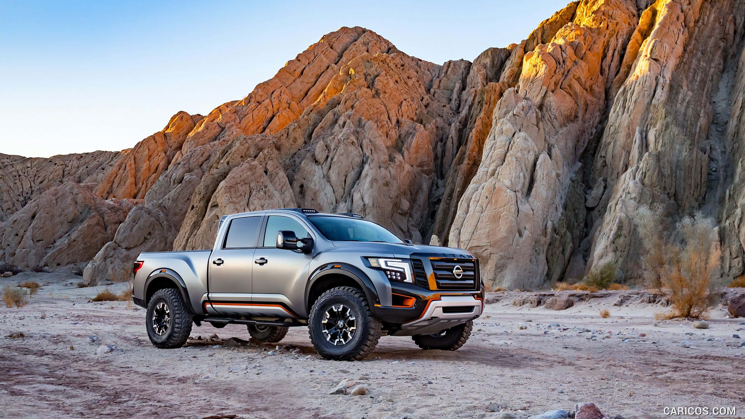 Nissan Titan 桌布 壁紙 — 一架銀色日產Frontier皮卡車矗立在崎嶇的沙漠地形上，戲劇性的岩石懸崖沐浴在金黃色的日落光芒中。