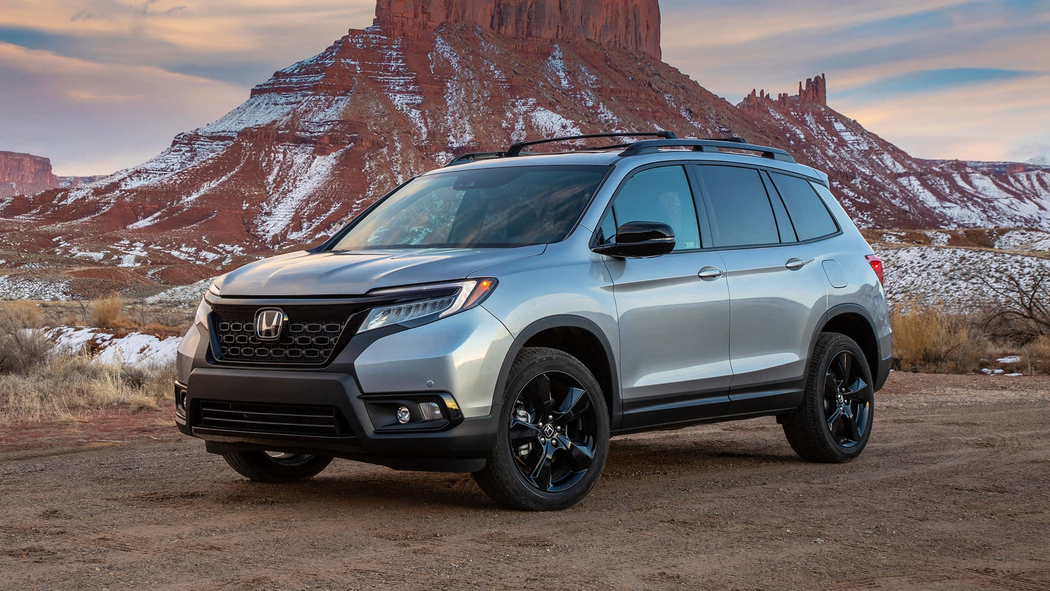 Honda Passport 桌布 壁紙 — 一架銀色本田Pilot SUV，配黑色輪圈，停泊在日落時分被雪覆蓋的紅色岩層前的泥路上。