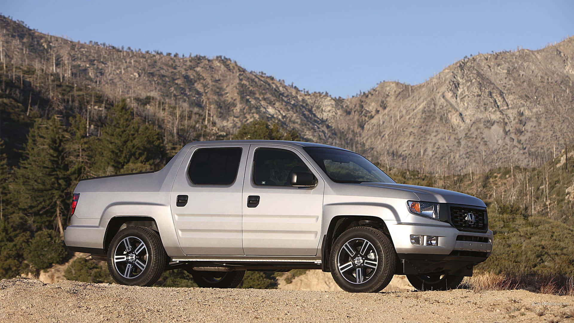 Honda Ridgeline 桌布 壁紙 — 一架銀色本田Ridgeline皮卡車停泊在泥路上，背景是崎嶇的山脈和常綠樹，天空清澈湛藍。