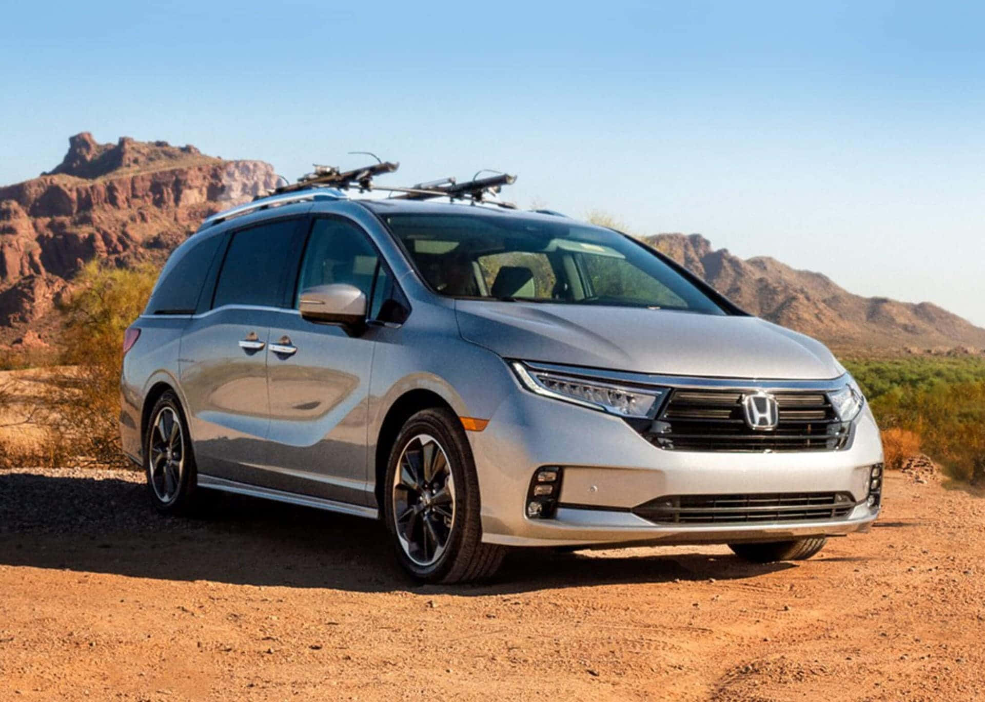 Honda Odyssey 桌布 壁紙 — 一架銀色本田小型貨車配備車頂行李架，停泊在塵土飛揚的沙漠道路上，背景是紅色岩石地貌，天空清澈湛藍。