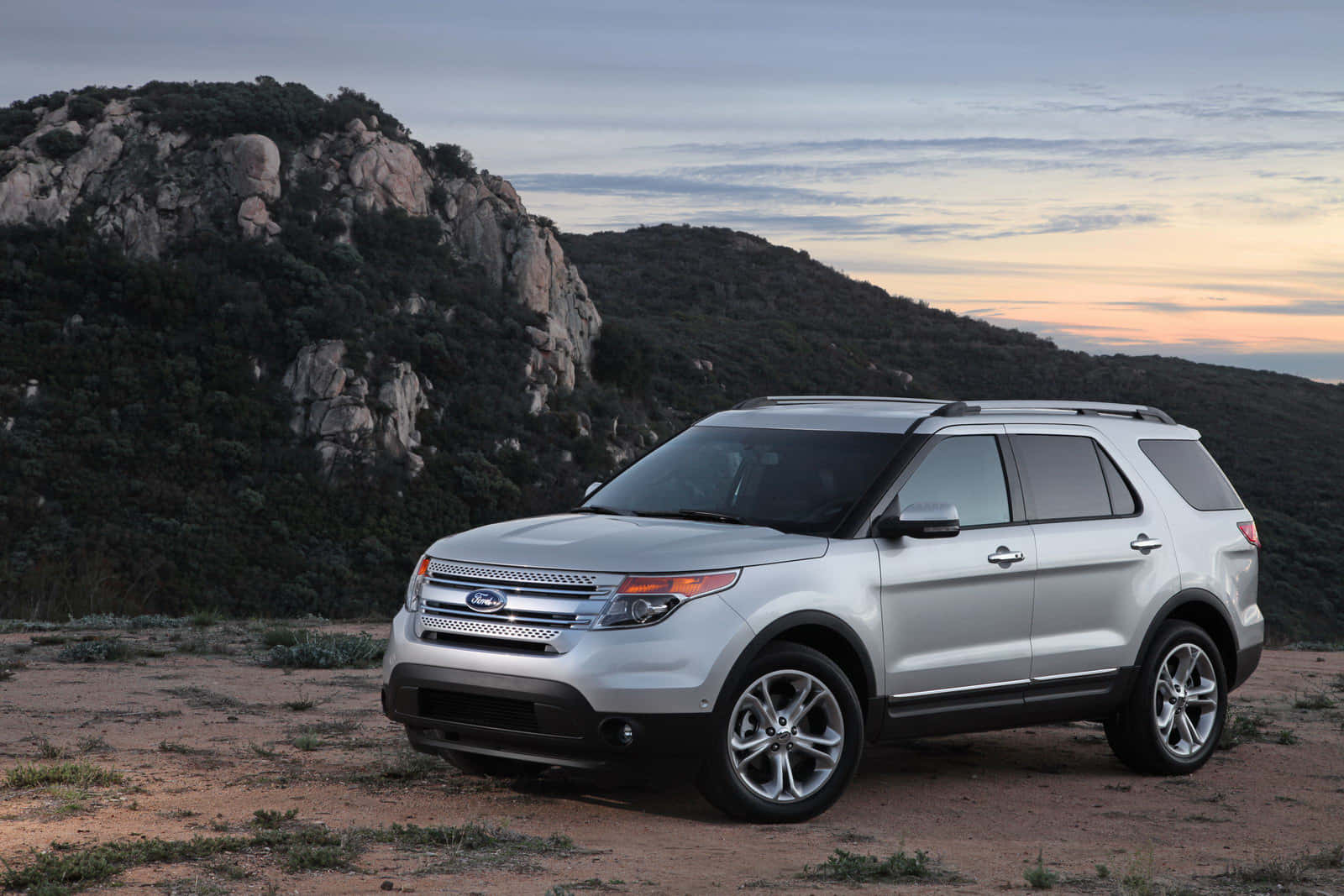 Ford Explorer 桌布 壁紙 — 一架銀色福特 Explorer SUV 停泊在塵土飛揚的地形上，背景有岩石丘陵和柔和嘅日落天空。