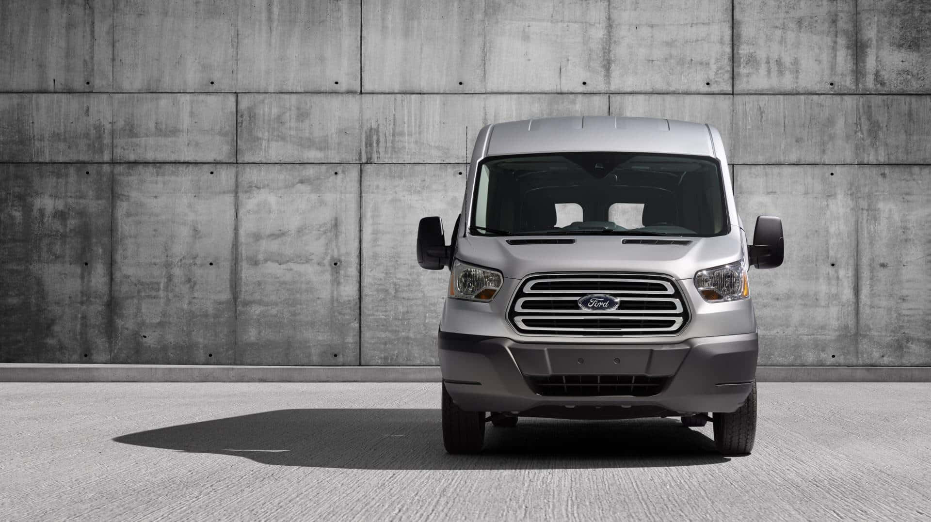 Ford Transit 桌布 壁紙 — 一架銀色福特 Transit 客貨車喺明亮日光下，面向一幅有紋理嘅混凝土牆。