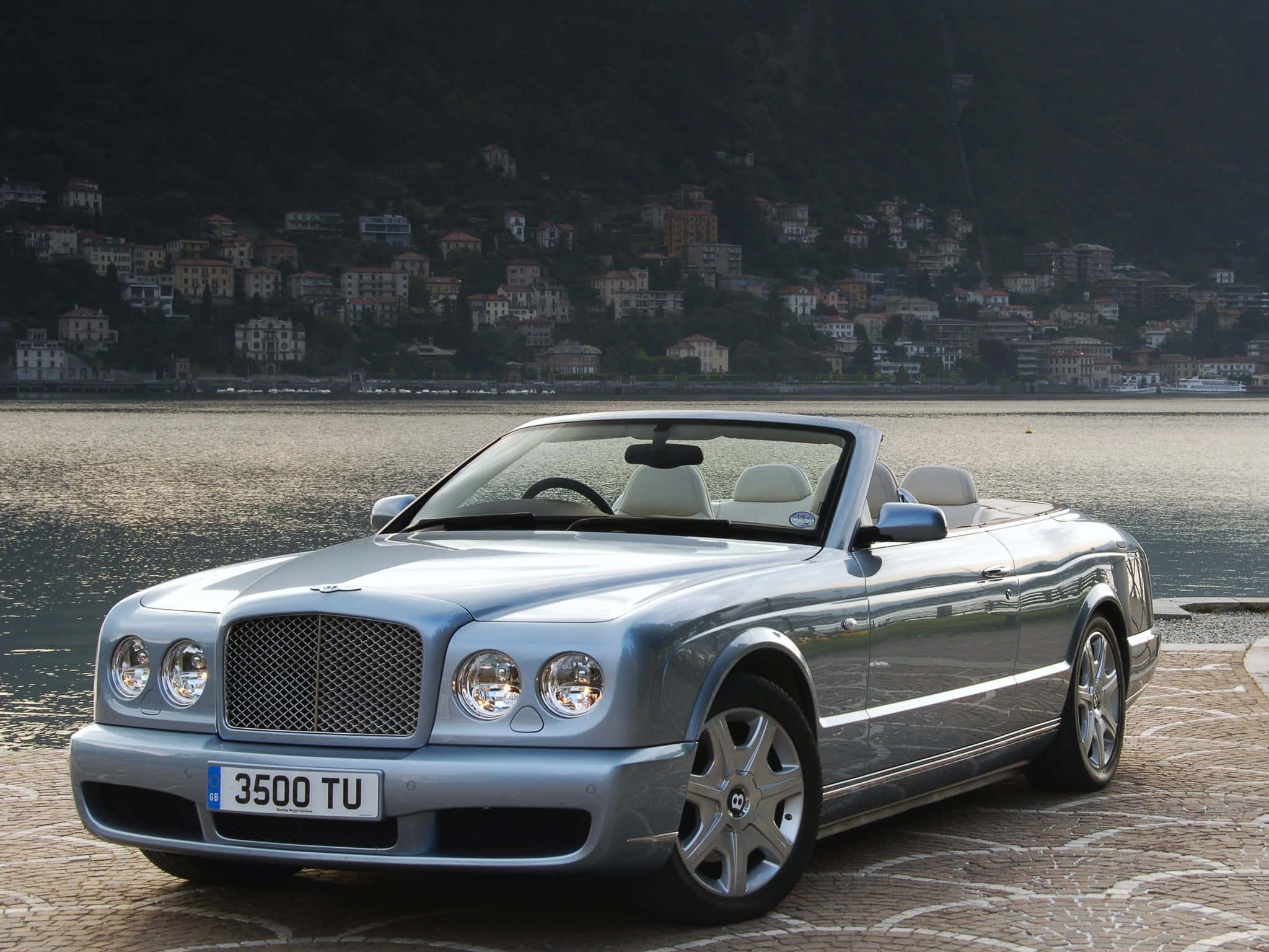 Bentley Azure 桌布 壁紙 — 一架銀色賓利開篷車停泊在平靜湖泊旁的石碼頭上，背景是山坡村莊。