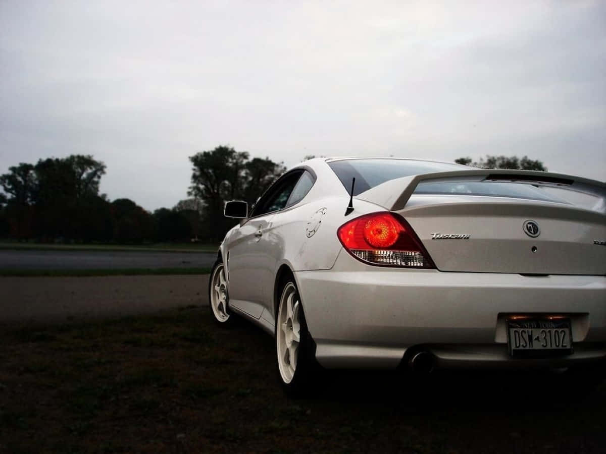 Hyundai Tiburon 桌布 壁紙 — 一架銀色跑車，紅色尾燈發光，停泊在路邊草地上，天空多雲。