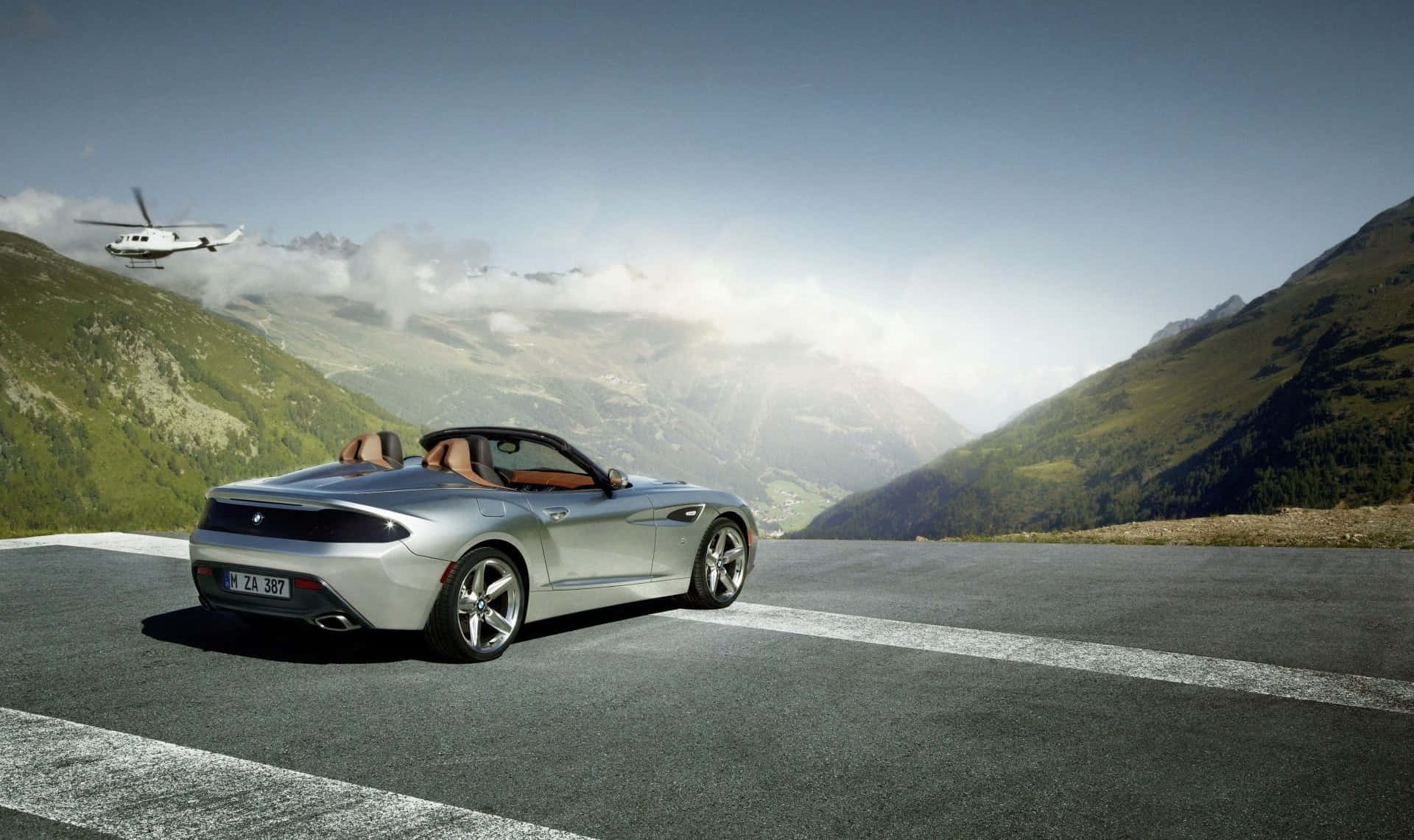 Bmw Z4 桌布 壁紙 — 一架銀色開篷跑車停泊在山路上，一架直升機在頭頂飛過，背景是綠色的山峰和雲層。