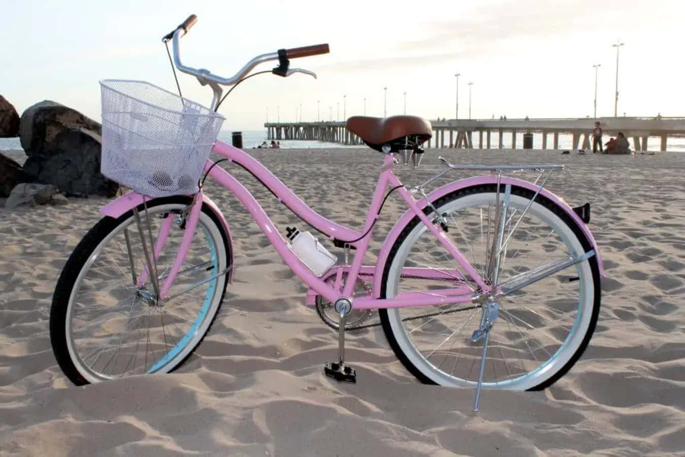 Beach Cruiser 桌布 壁紙 — 一架閃亮的粉紅色沙灘巡航自行車，配有鐵絲籃，停泊在日落時分碼頭附近的沙灘上。